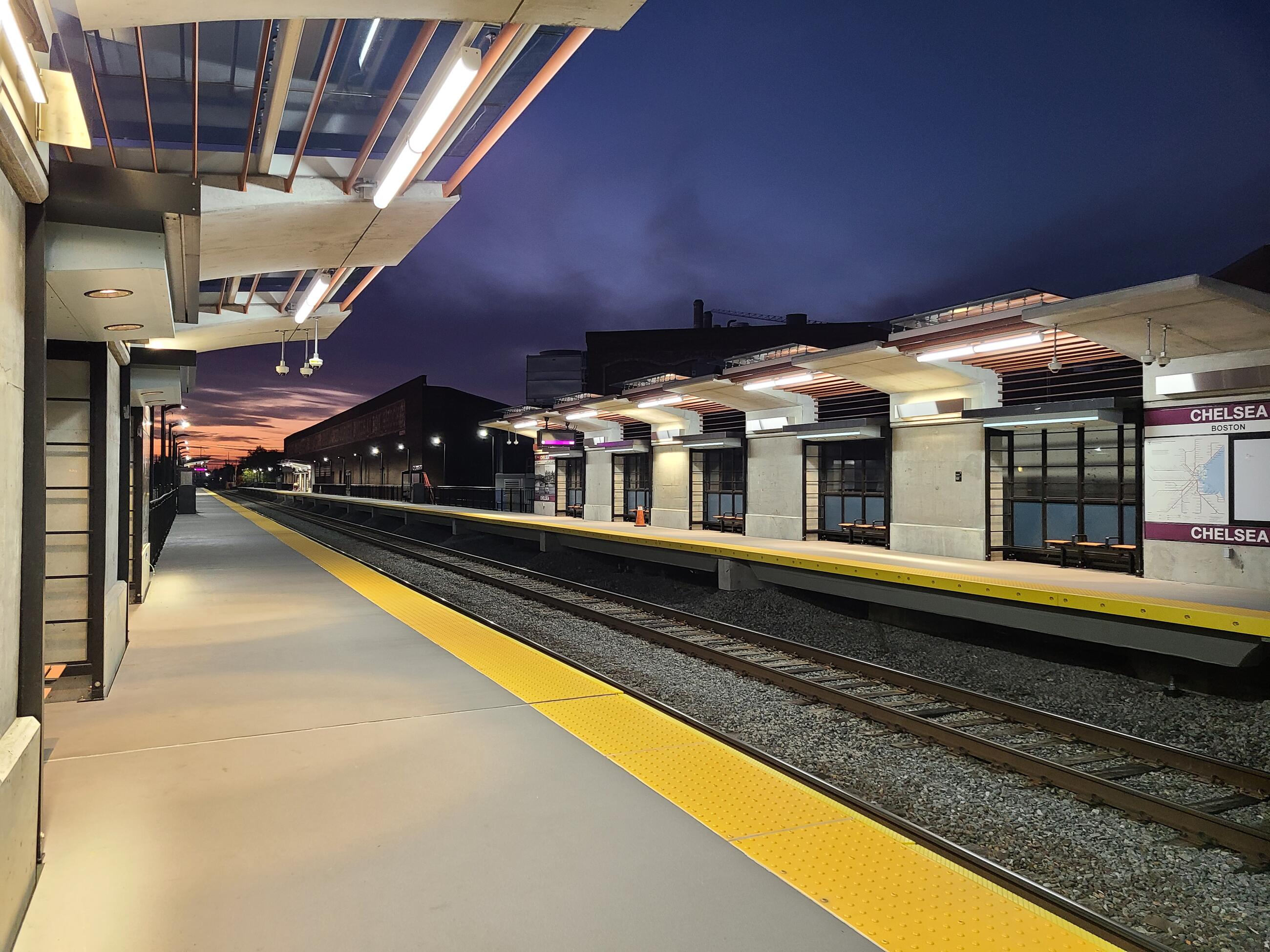 The new Chelsea Commuter Rail station is shown at dusk. The overhead lights that hang from the clear canopies over the platform are on. They illuminate the platforms at the left and right sides of the image, with the empty track running down the middle. The large Chelsea Station sign and map are shown along the right platform, and there are benches in the recessed lighted areas under the canopies. 