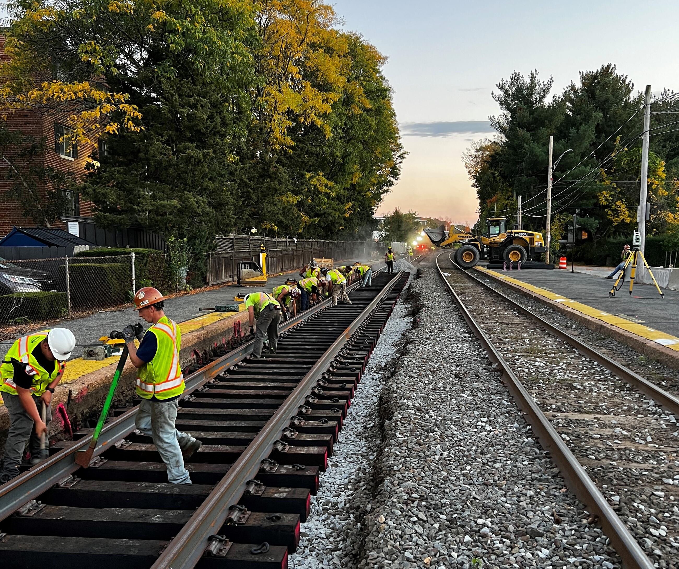 Ten crew members in yellow vests and hardhats work in a line along Track 1 replacing rails and ballasts on Track 1 at Melrose/Cedar Park station. 