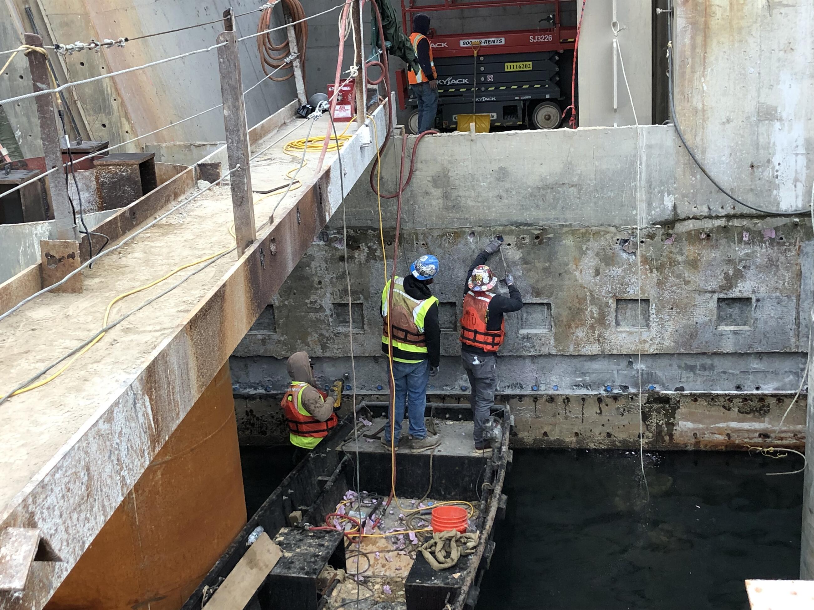 Three workers in safety vests stand on a small wooden boat on the water next to the bridge. They are cleaning the formwork from the bridge's south footing.