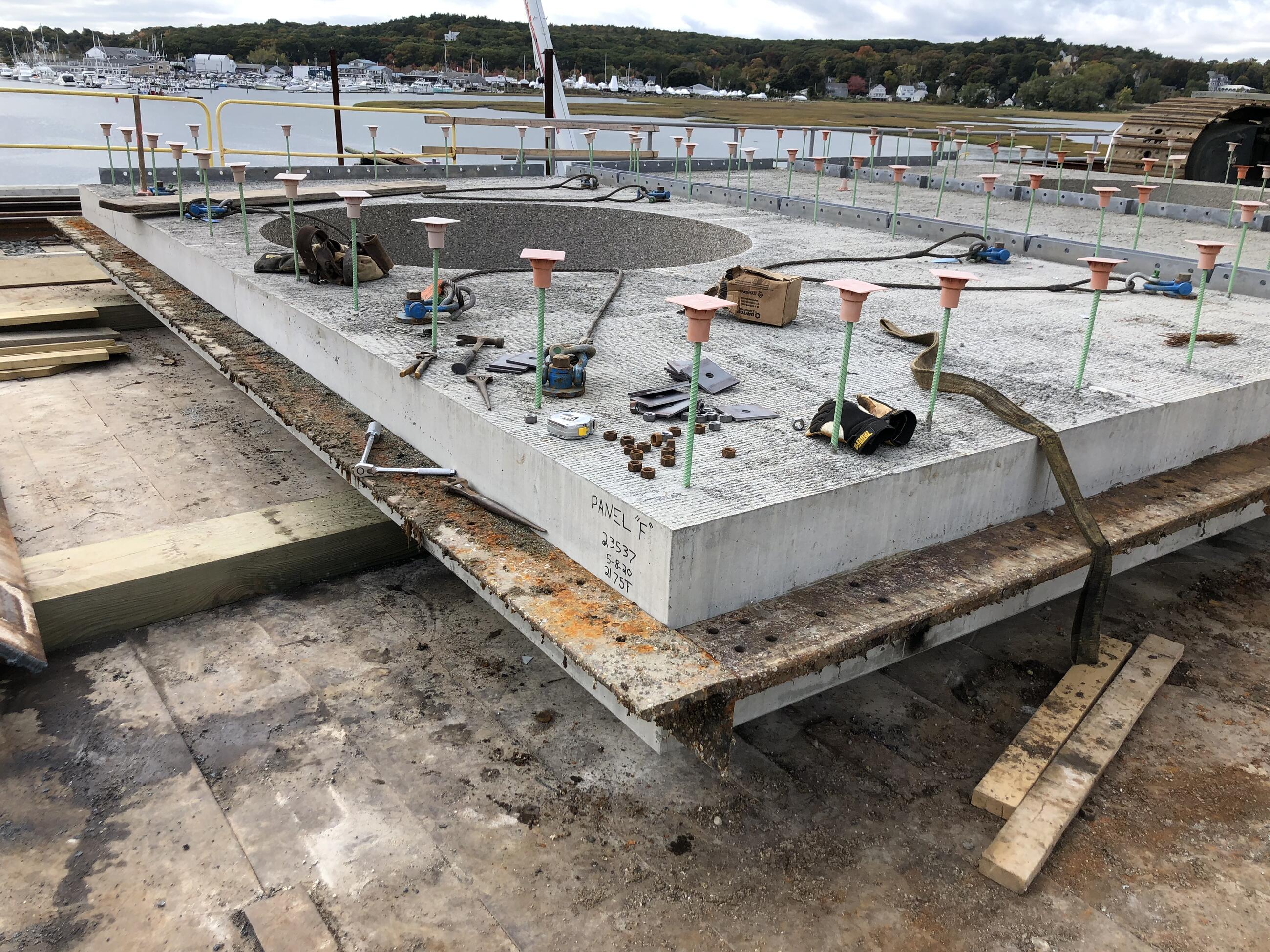 A large, rectangular, poured-concrete footing panel is shown resting on a plywood structure near the water before placement and installation. 