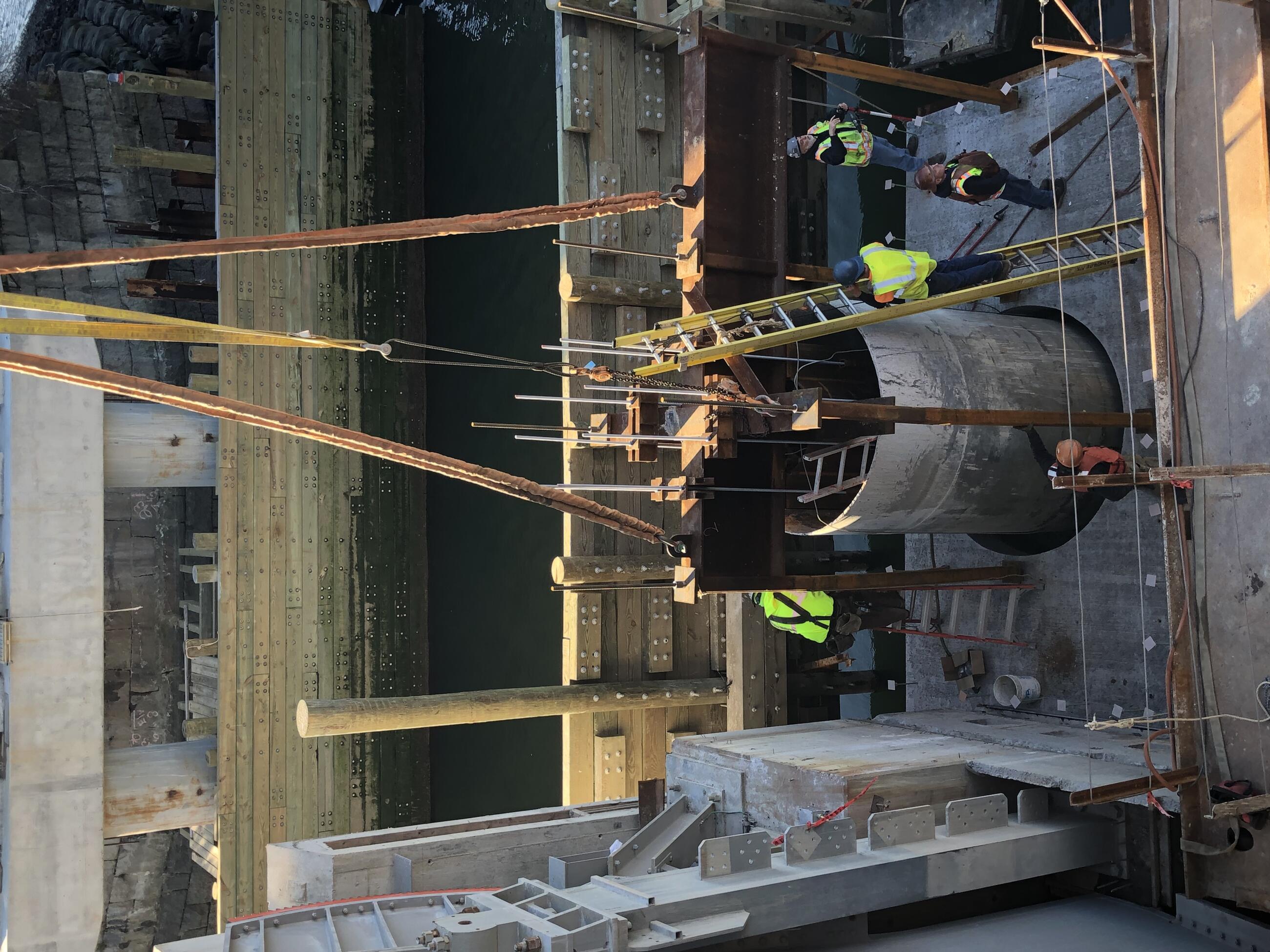 Five workers stand on (or on ladders resting on) a precast, poured-cement footing panel just after it has been lowered into place. Suspension cables are still attached to the footing.