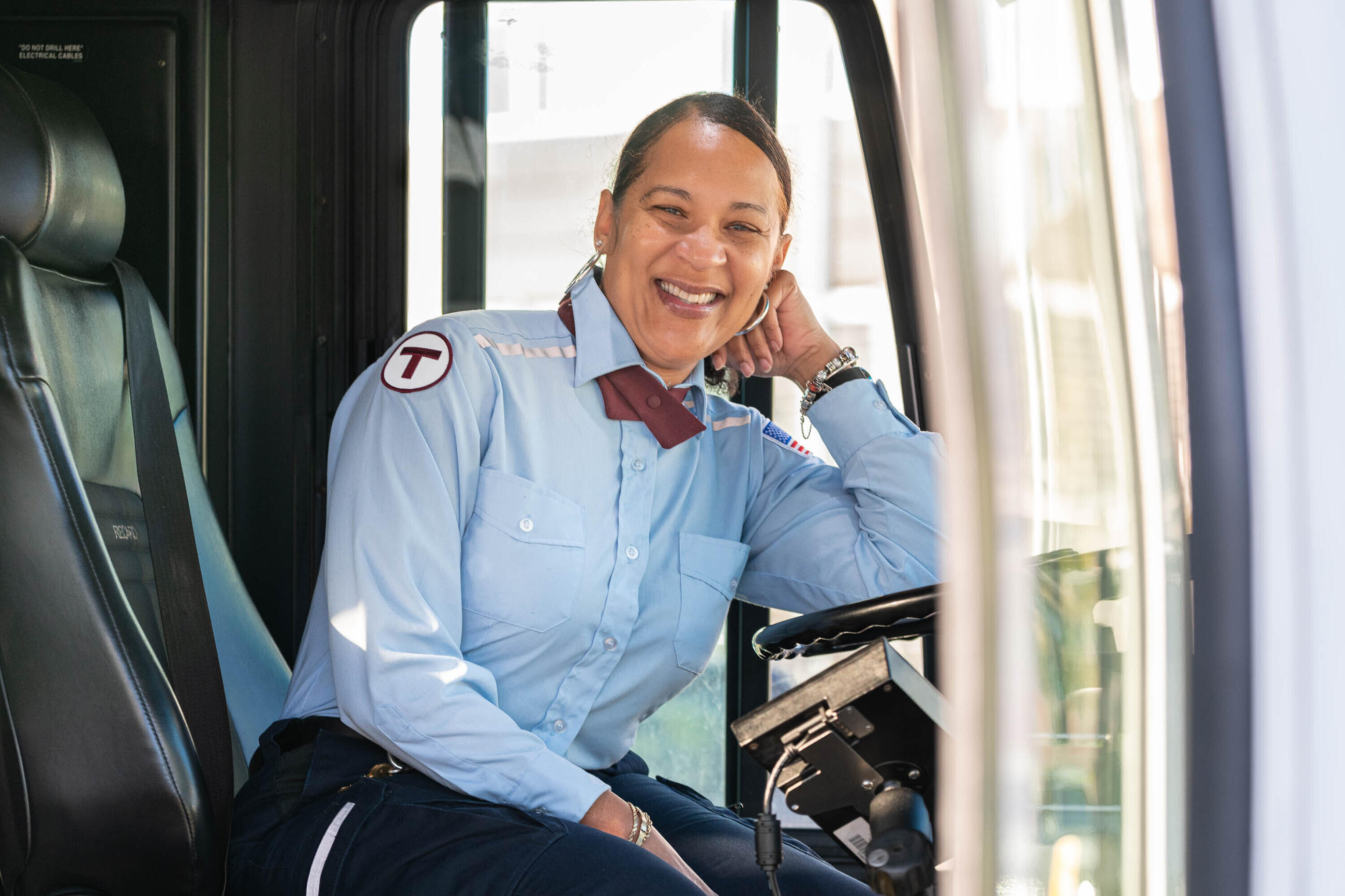 A bus operator in uniform smiling with her elbow resting on the wheel and her hand by her face
