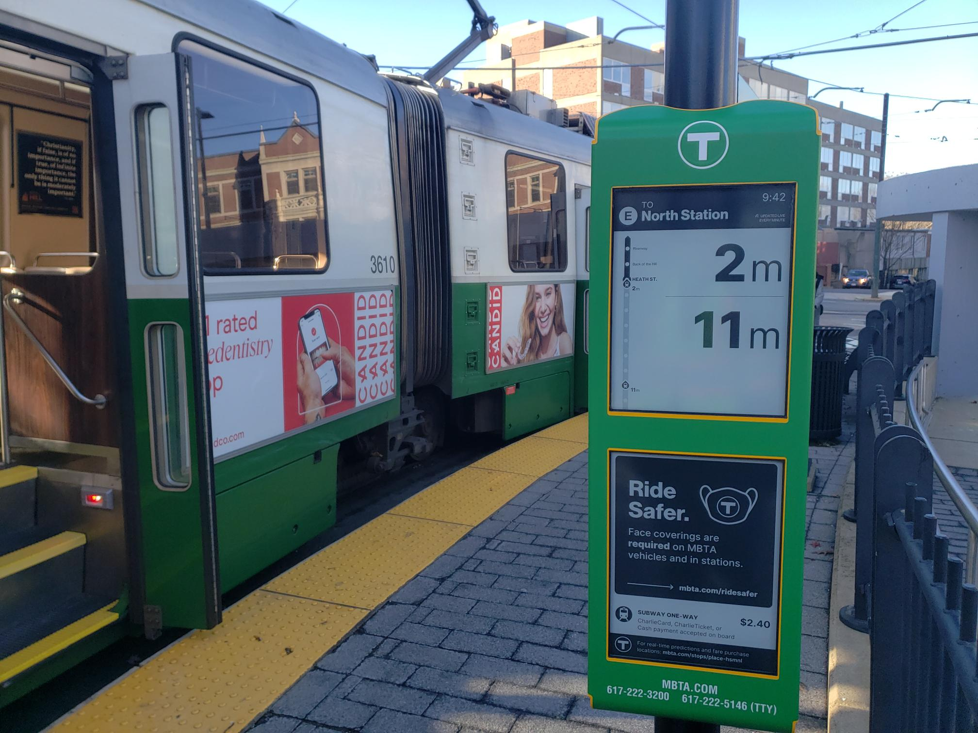 A photo of an e-ink sign at Heath St with a green line trolley waiting with open doors next to the curb