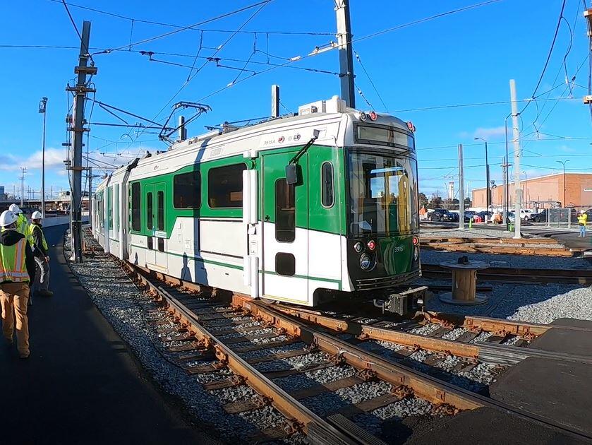 A green and white Green Line Extension test trolley travels on tracks as crew members stand nearby and observe. The sky is bright blue on a clear December morning, and the train casts shadows across the tracks. 