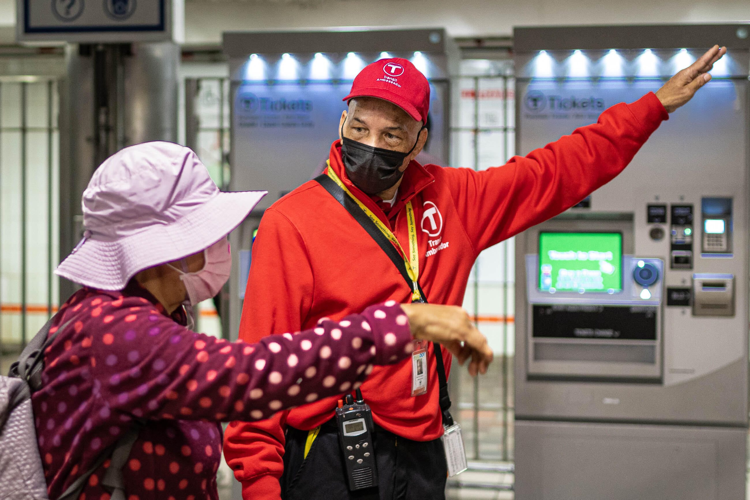 A transit ambassador stands inside a station in front of fare purchase machines and provides an elder with directions. The two people face each other and both point to the right.