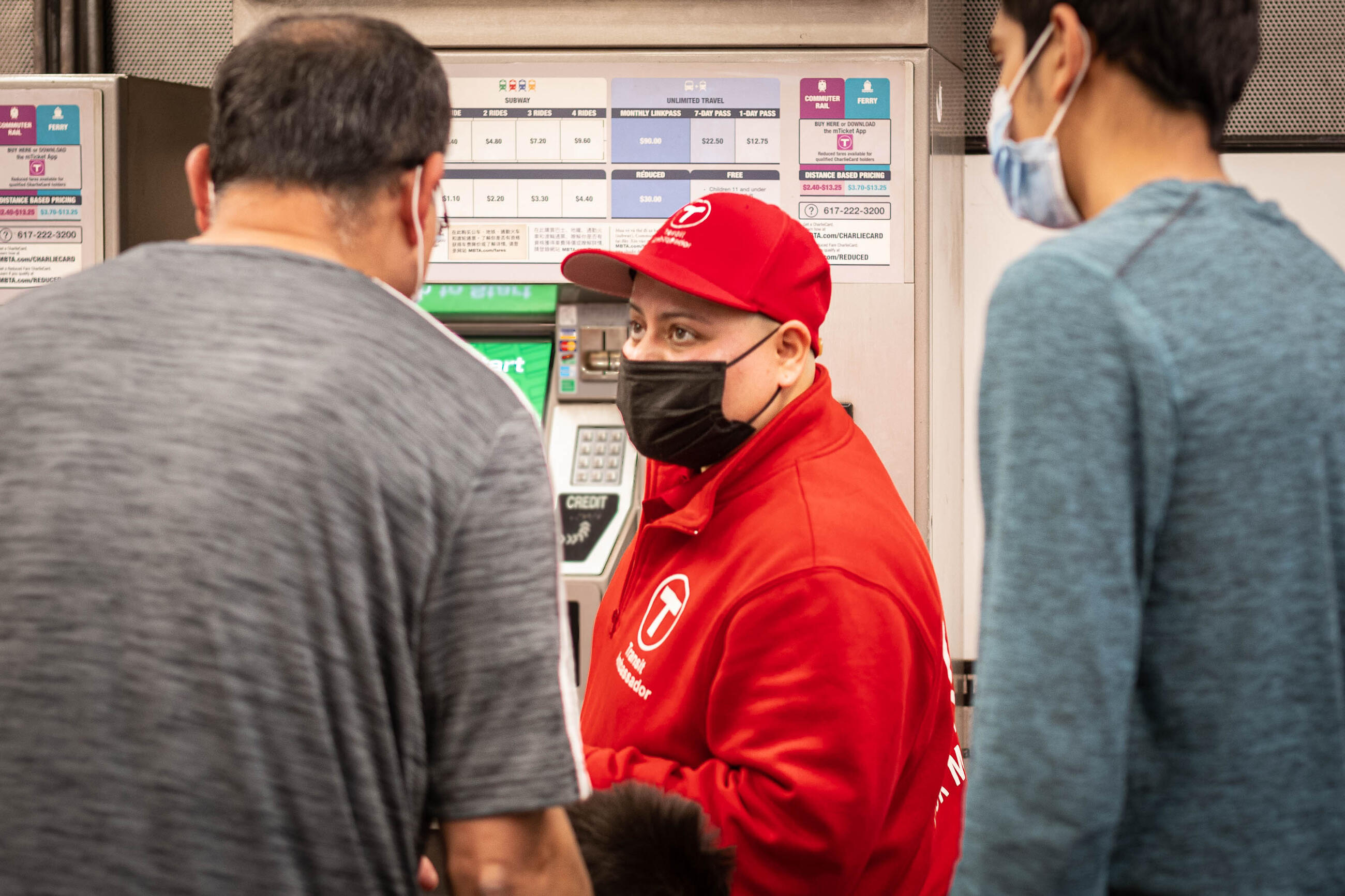 A transit ambassador in a red shirt and cap faces the camera and while two riders face away, toward the ambassador. They all stand in a station in front of fare machines. 