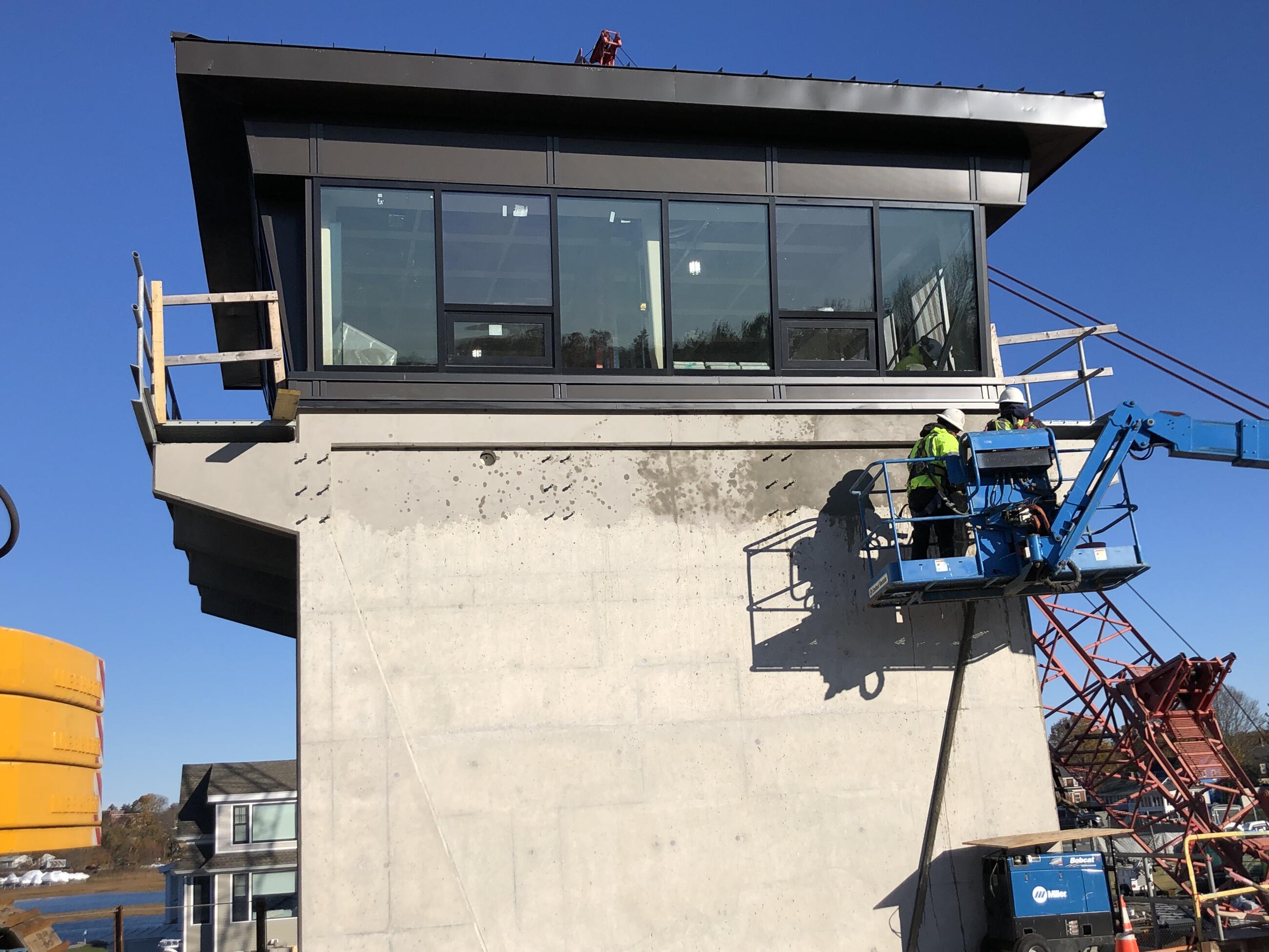 Two workers in a cherry picker work on the exterior of the control tower. Another worker can be seen through the recently installed windows working inside the control tower.