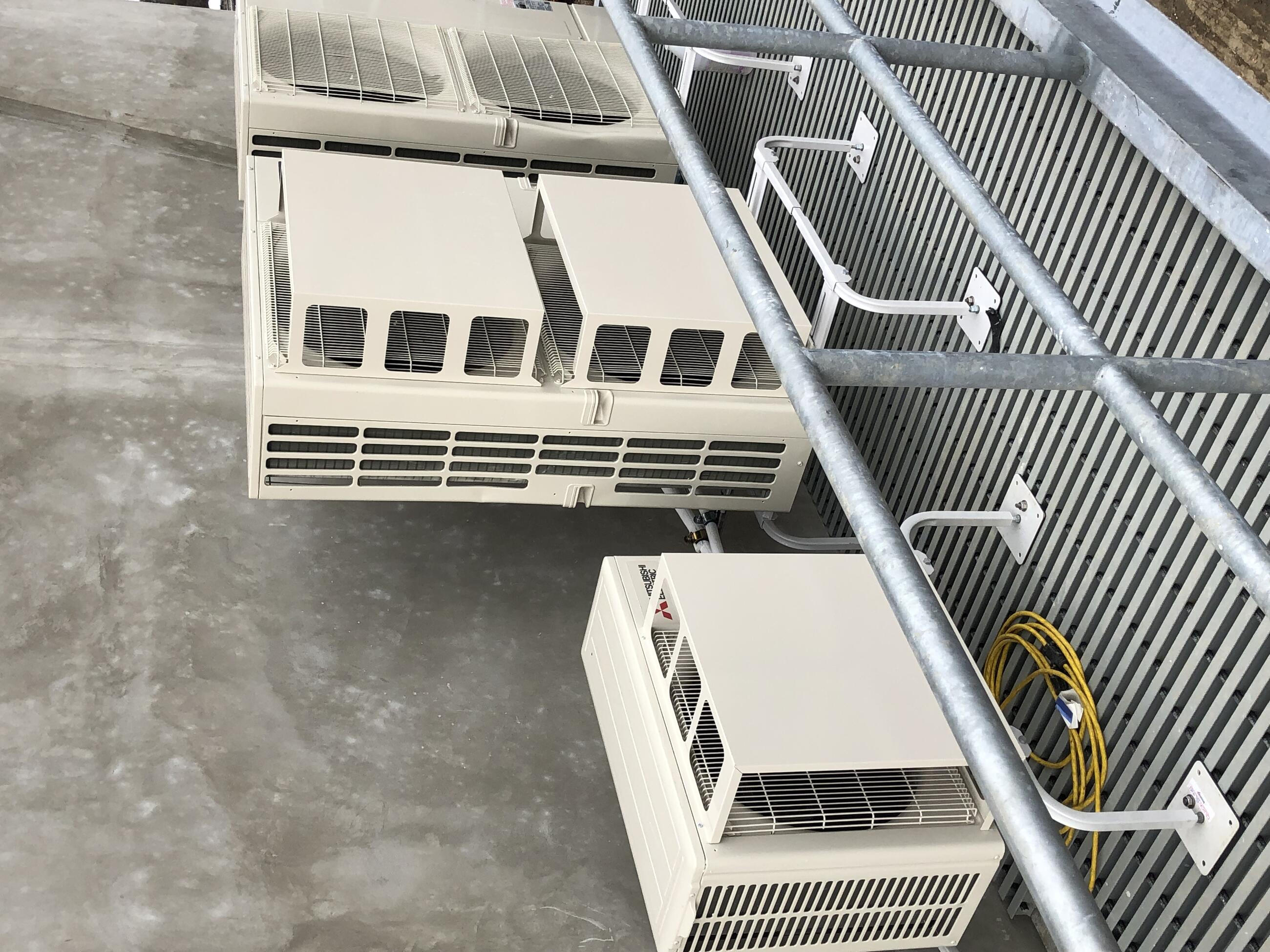 Three HVAC units are shown installed over a metal grate next to a cement wall. The units are bolted to metal legs that lift them off the grate to allow for improved airflow.