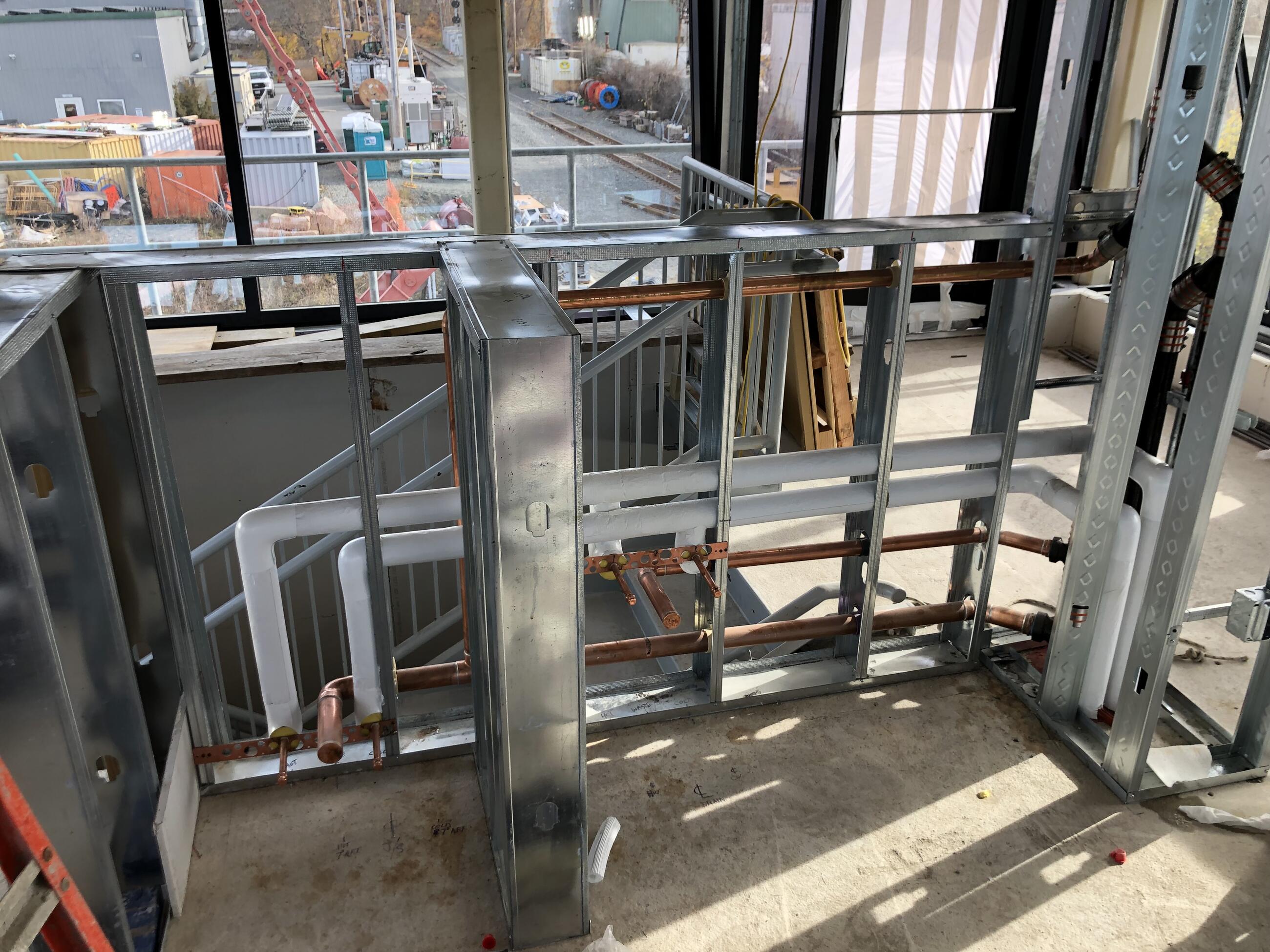 Image shows the metal structure where the control tower's kitchenette will be installed. Copper plumbing pipes have been installed within the metal structure. The view faces a window overlooking construction materials and train tracks. 