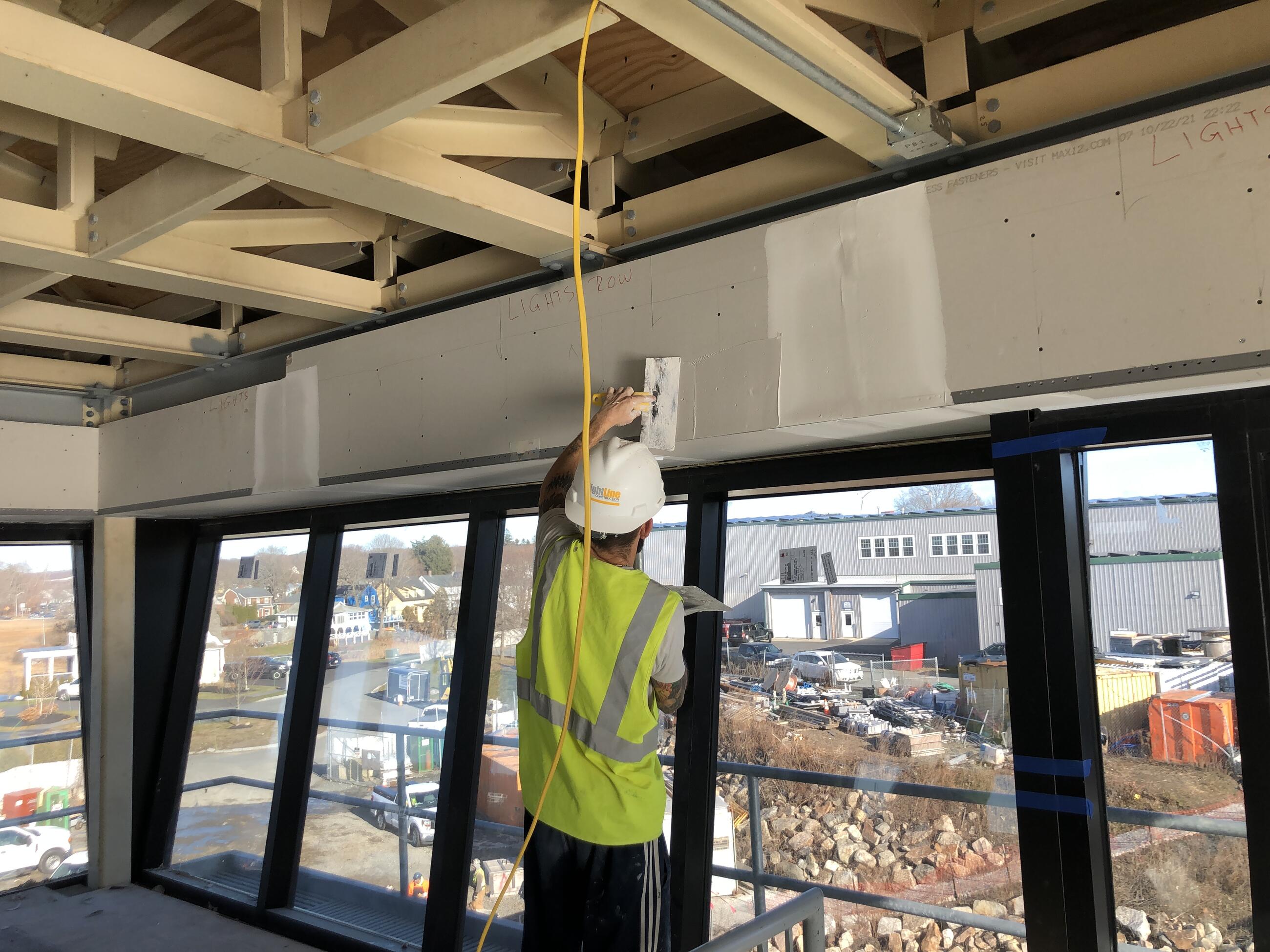 A worker in a yellow vest and hardhat smooths plaster over the joins in a span of drywall above windows in the control tower. The windows overlook the construction site below. The wooden beams below the ceiling of the control tower can be seen above the worker's head. 