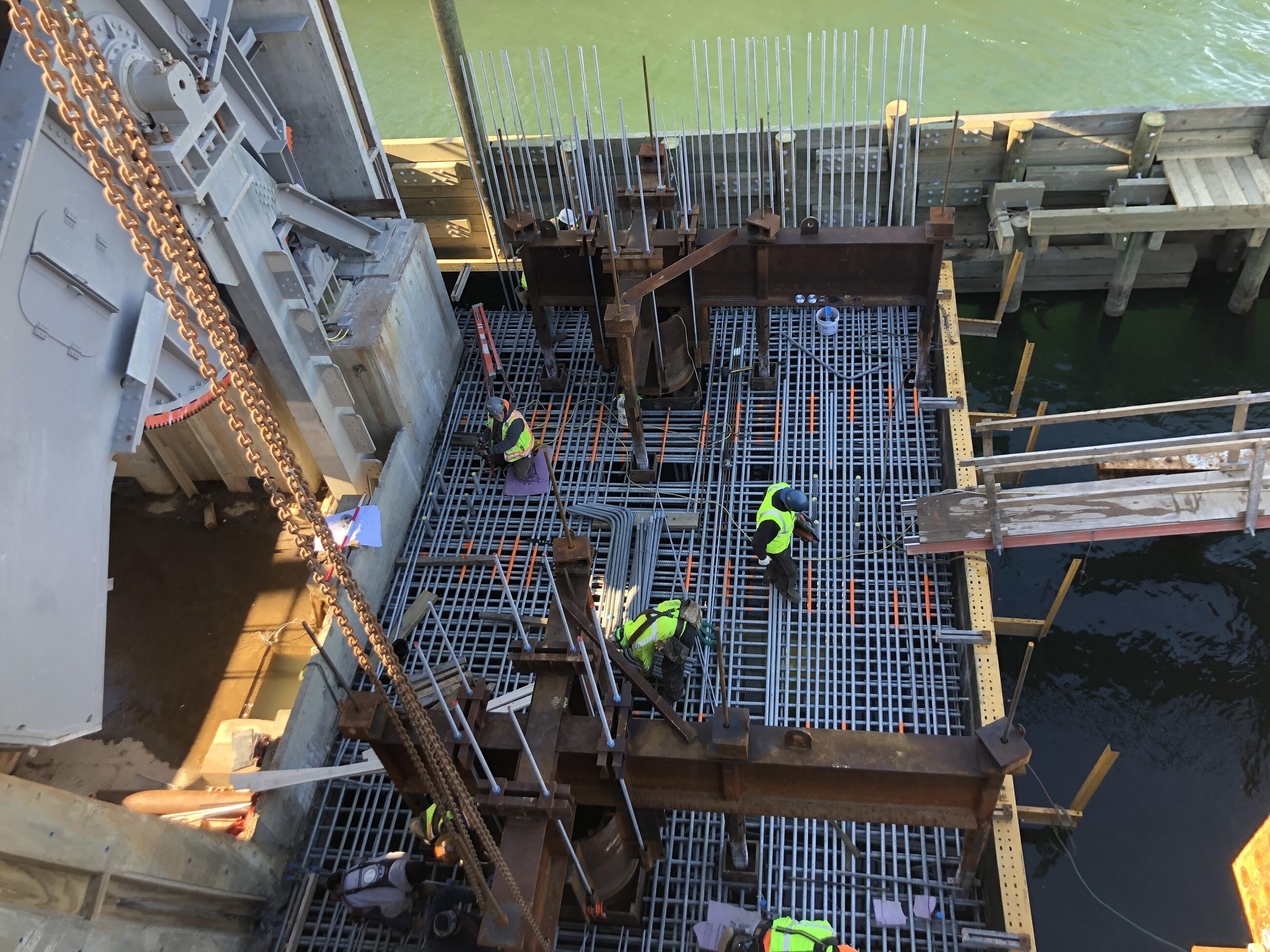 In an image photographed from overhead, four workers create the reinforcing grid of metal bars that will be embedded within the poured concrete to form the pier cap. They stand and kneel atop the gridwork before the concrete is poured. The movable bascule span is shown at left, the waterway at the top of the photo.