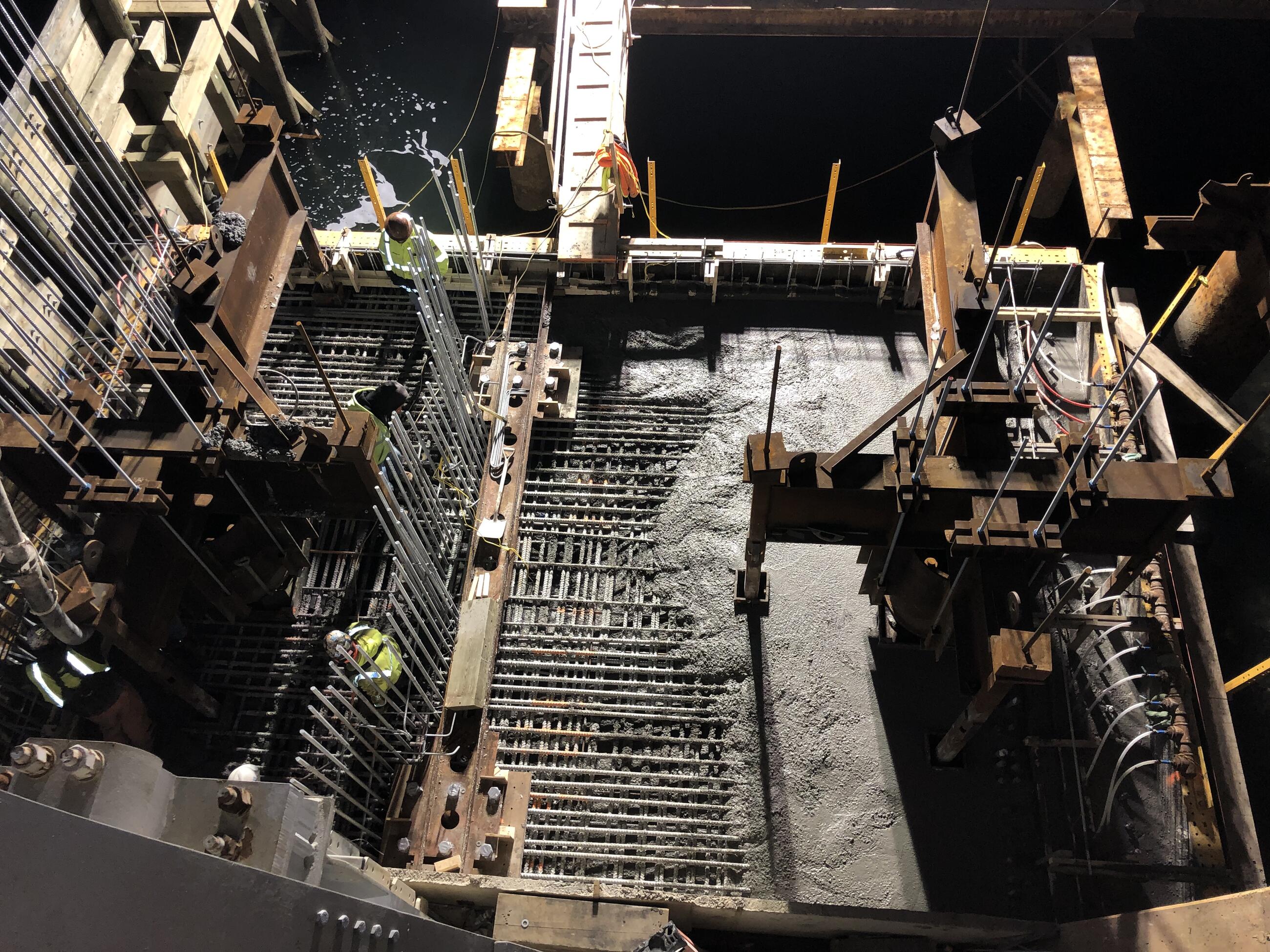 An overhead view of cement being poured into a large form to create the pier cap. Three workers stand at the left edge of the form on top of a gridwork of rebar that will be embedded within the concrete. 
