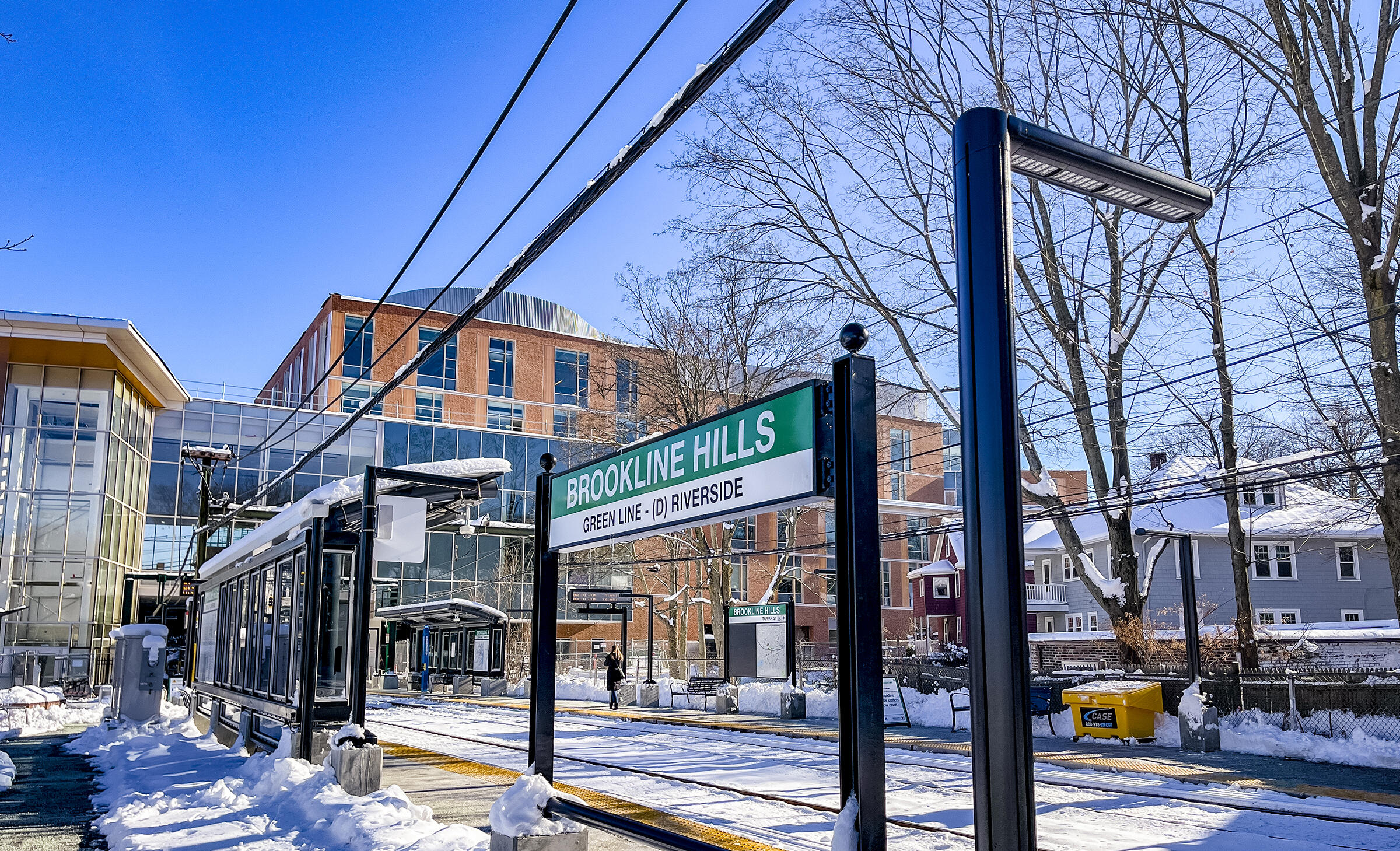 A photo of the Brookline Hills station sign in front of the platform on a bring sunny day with snow on the ground 