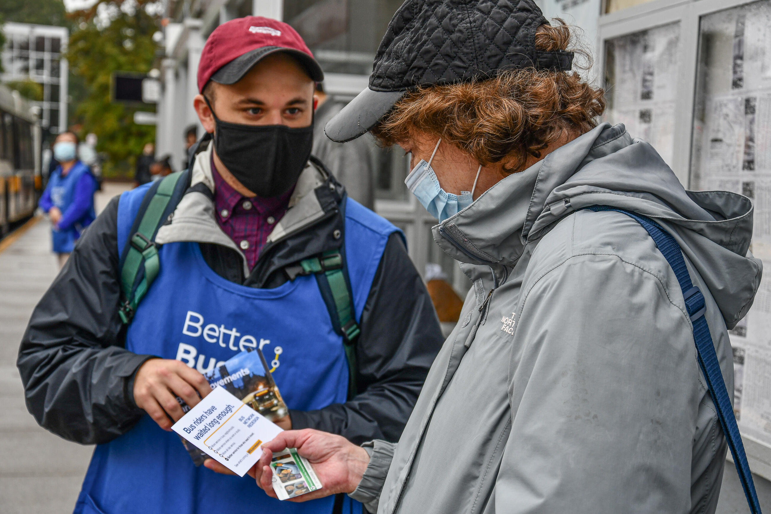 A member of the MBTA's System-Wide Accessibility team wearing a red hat, mask, and an apron that says Better Bus Project on the front, talks with a rider, also in a hat and mask, about the Bus Network Redesign project as they two stand outside at Quincy Center. 