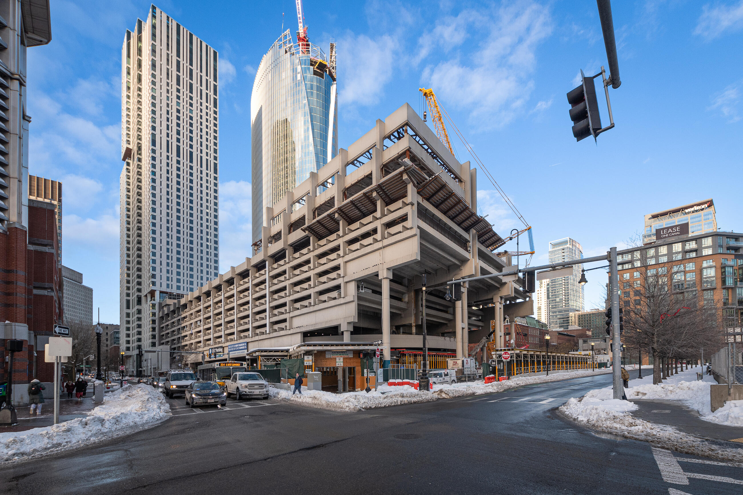 The Government Center Garage is shown at center with cross streets in front and tall buildings behind. Two large construction cranes sit atop the garage, and another is at street level in front of the garage at right.  