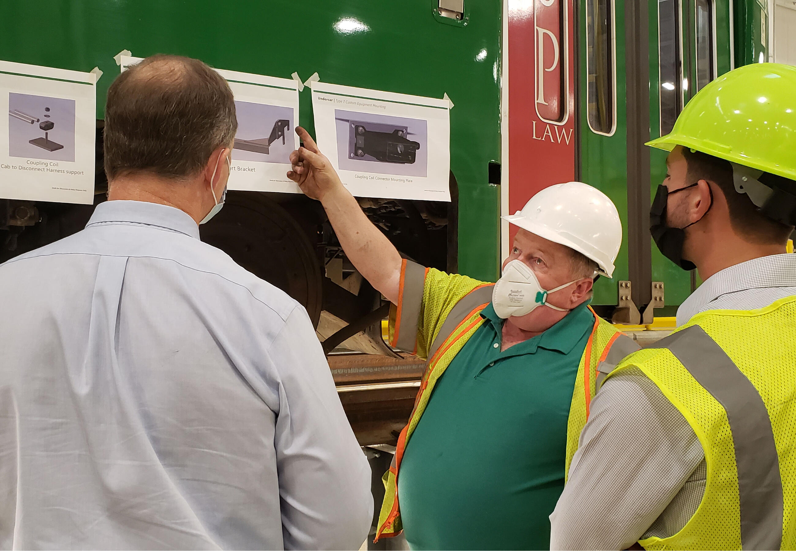 A man in a hard hat points to equipment photos taped on the outside of a green line trolley while two other men watch him give an explanation