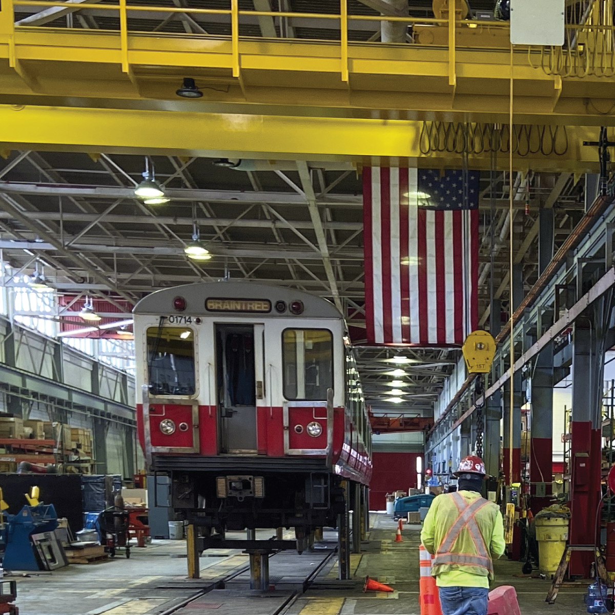 a red line train car on a lift in the cabot maintenance facility with an american flag hanging from the ceiling nearby
