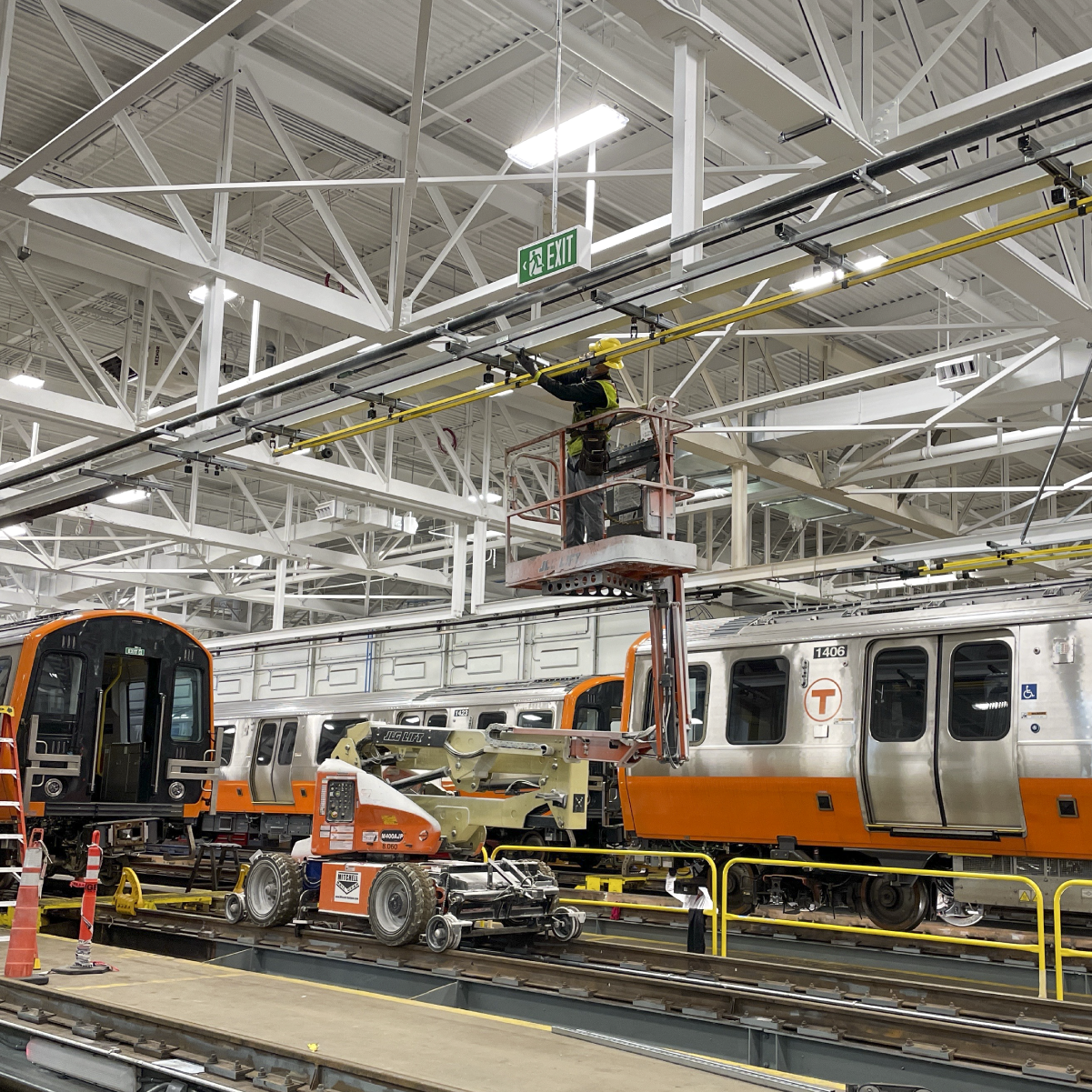 worker in a lift installing equipment inside the wellington facility with new orange line trains on the maintenance tracks in front of and next to them 