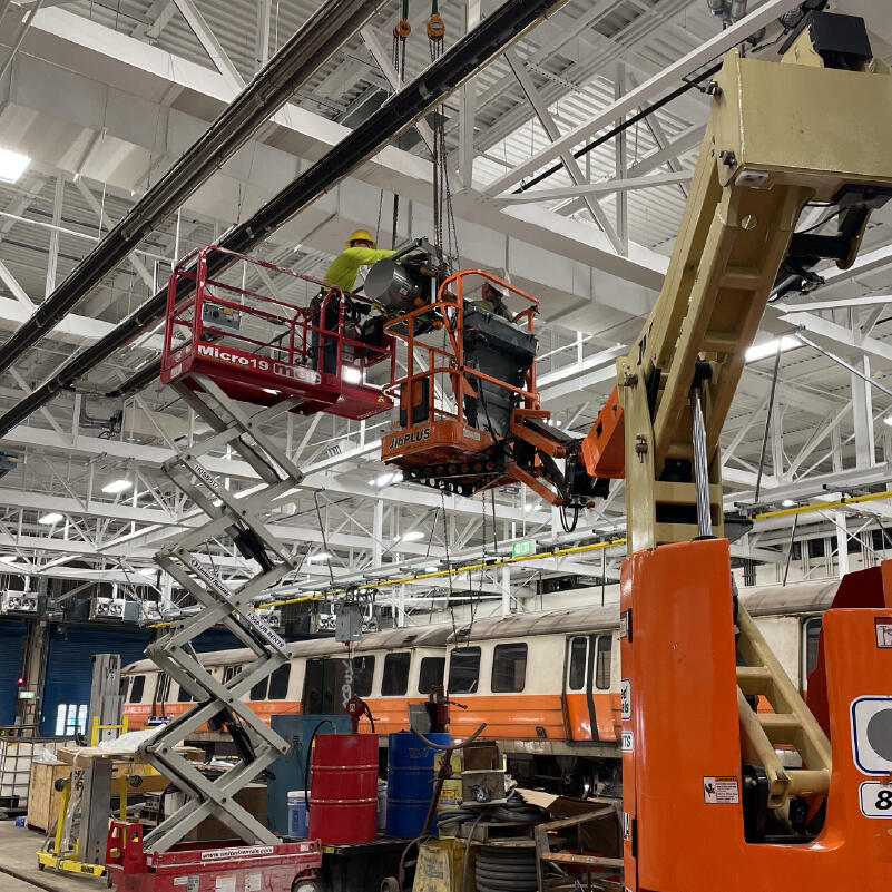 crew members working in lifts inside the wellington vehicle facility with an orange line train visible in the background