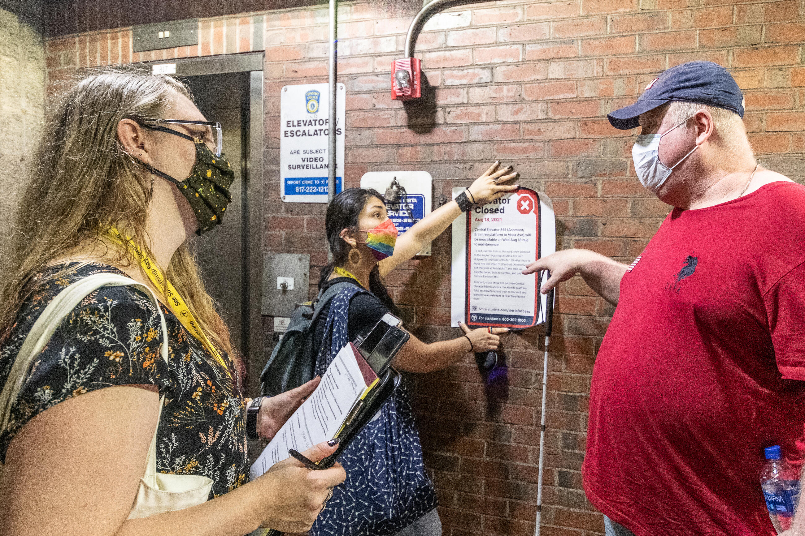 An elevator-dependent rider holding a white cane talks to a researcher with a clipboard and another who is holding up an "ELEVATOR CLOSED" sign next to an elevator at Davis Station. 