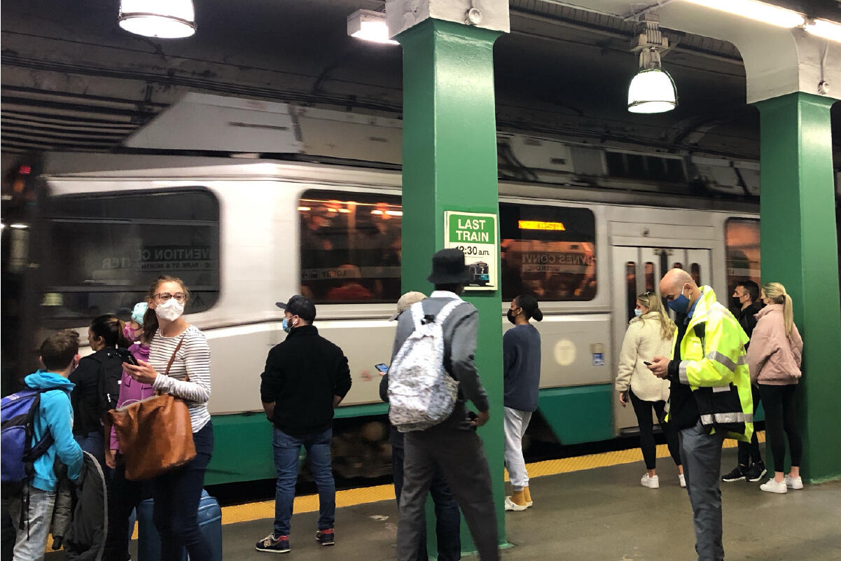 many riders wearing face masks stand on the platform as a green line train pulls up inside the station