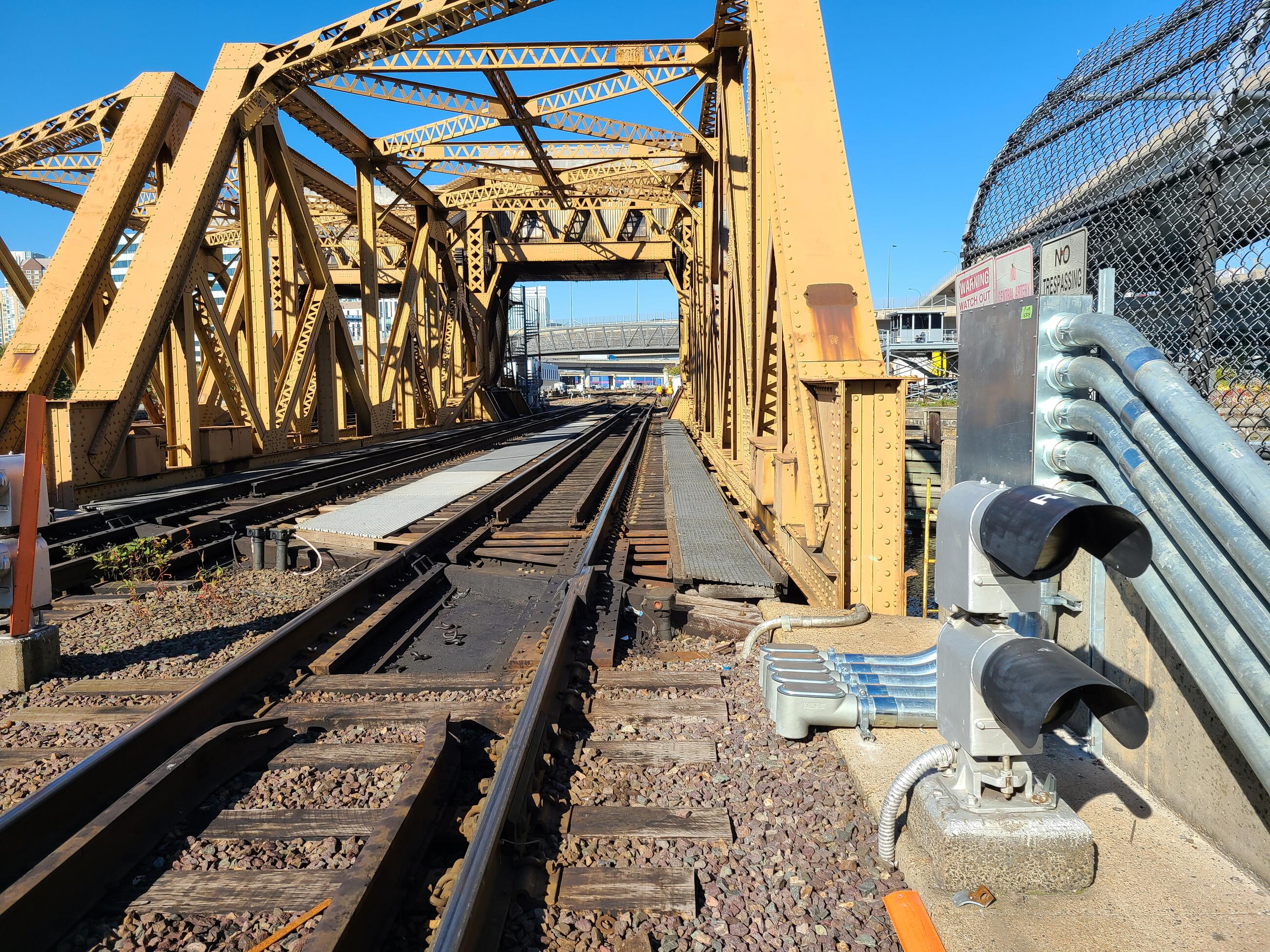 A yellow-painted riveted steel drawbridge hovers over an empty track at a North Station drawbridge. Signal lights with curved black metal over their tops shielding them from direct sunlight are next to the bridge at right.