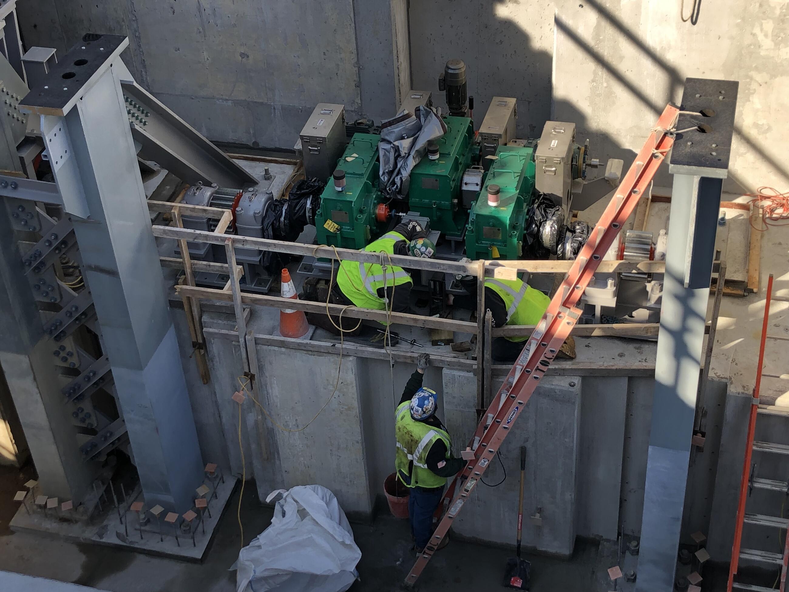 Three workers in yellow vests install machinery above a poured concrete structure, with steel posts pointing skyward on either side of them. Plywood temporary railings run along the side of the platform on which they work, and tall ladders lead from the base of the pier to the work area.
