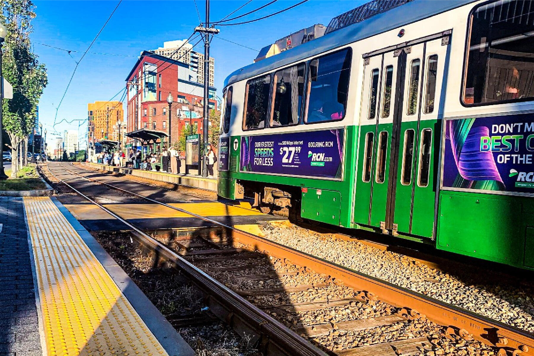 a green line train approaching an e branch station platform where riders are waiting to board