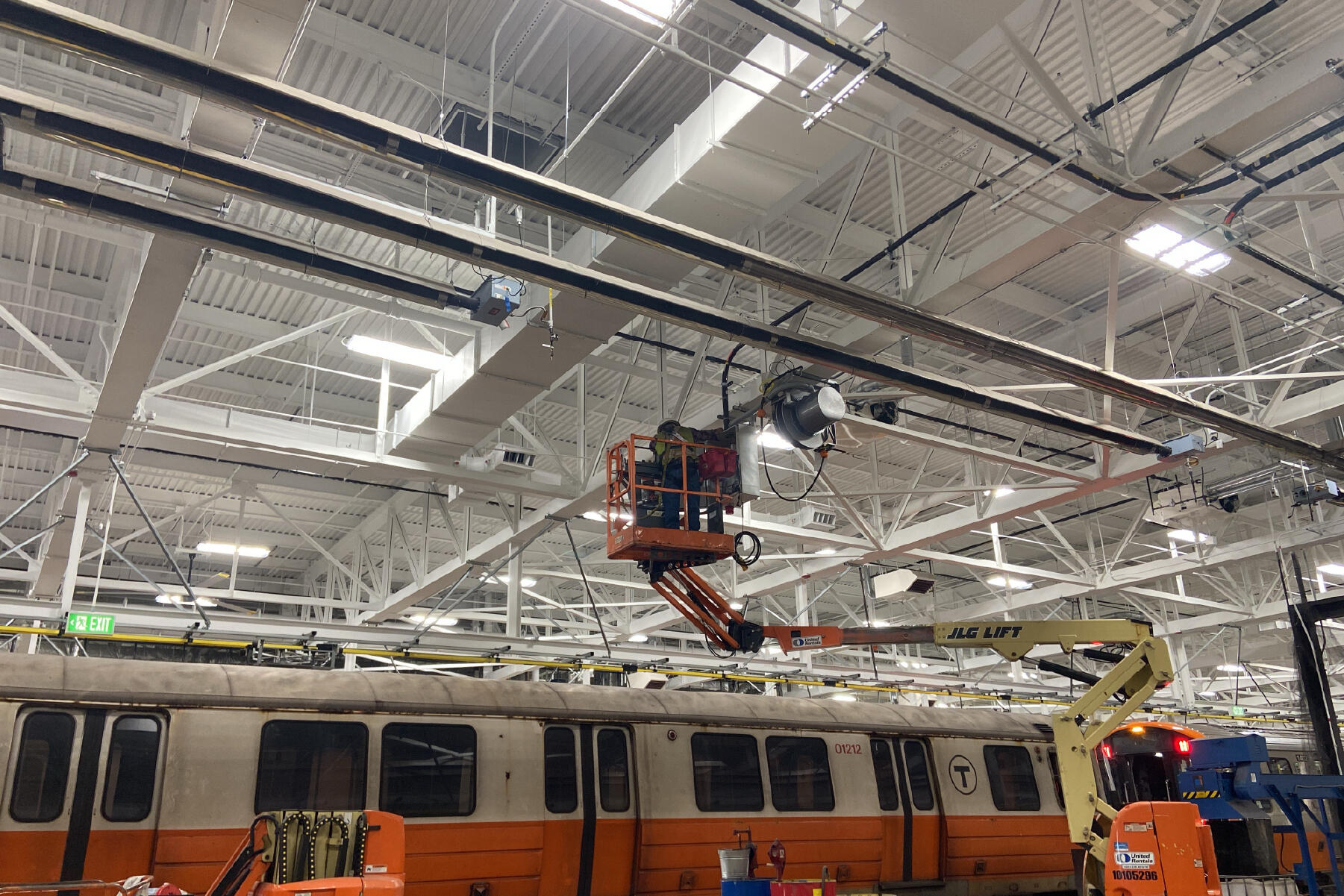 a crew member in a lift working on something near the ceiling above an old orange line car inside the maintenance facility
