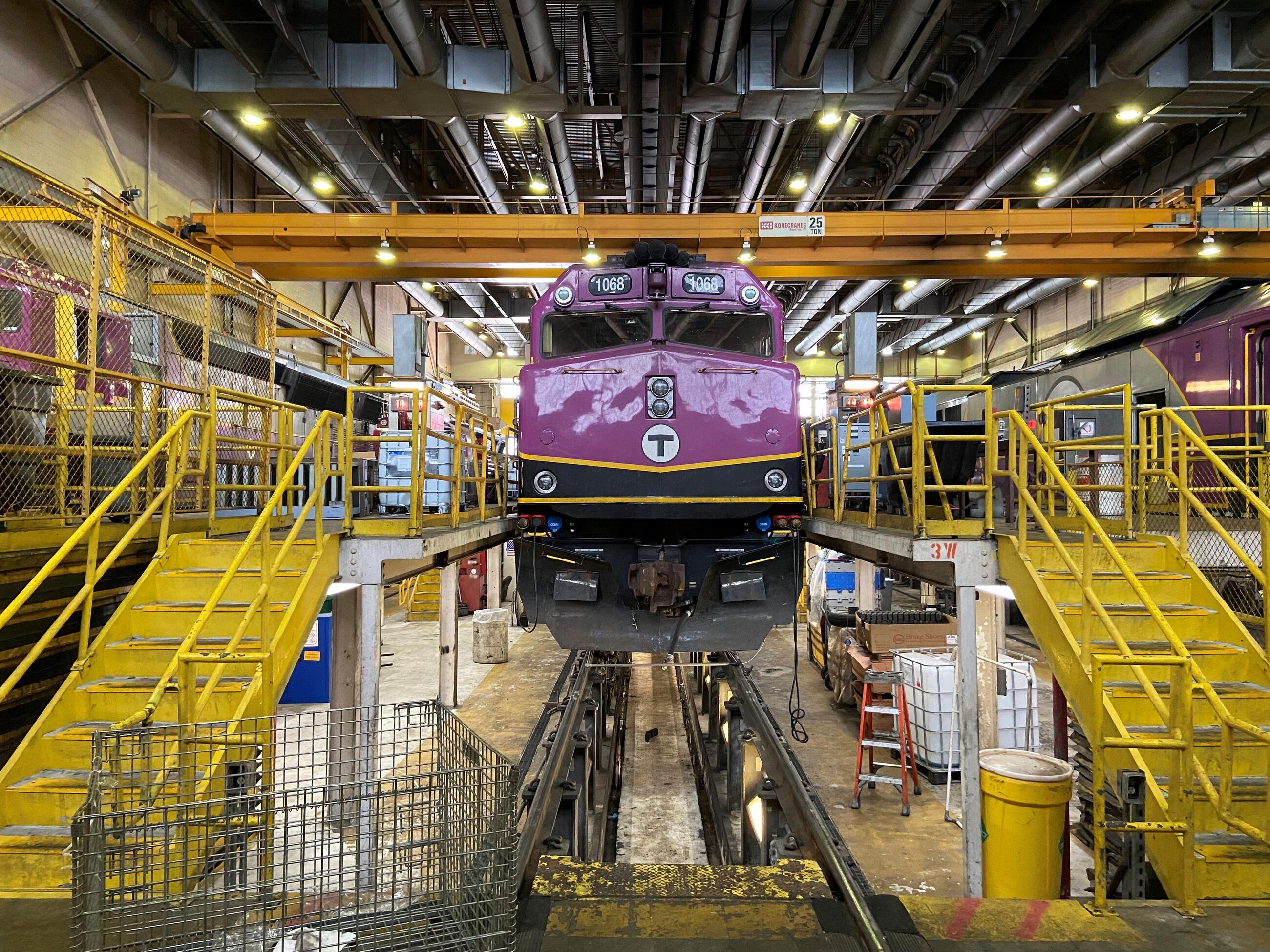 Commuter Rail train 1068 is shown in the center of a busy scene from the Boston Engine Terminal maintenance facility. It is up on high tracks that allow workers to work on the train from below. Yellow metal stairs and railings on either side of the train allow workers to enter the train from above. Many silver ducts and lights right overhead, and more lighting from below the train illuminates it from underneath.