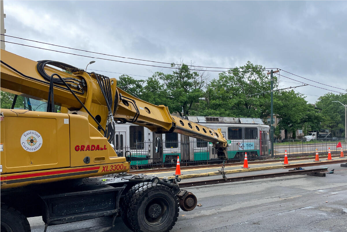 A hydraulic excavator holds a new rail that is being prepared for installation on the B Branch. A green line train passes by in the background.