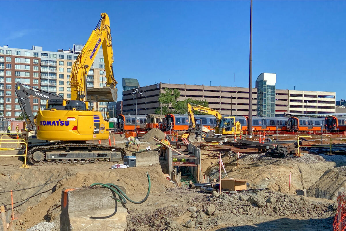Workers construct duct banks at Wellington Yard. Construction equipment and orange line trains are parked in the background