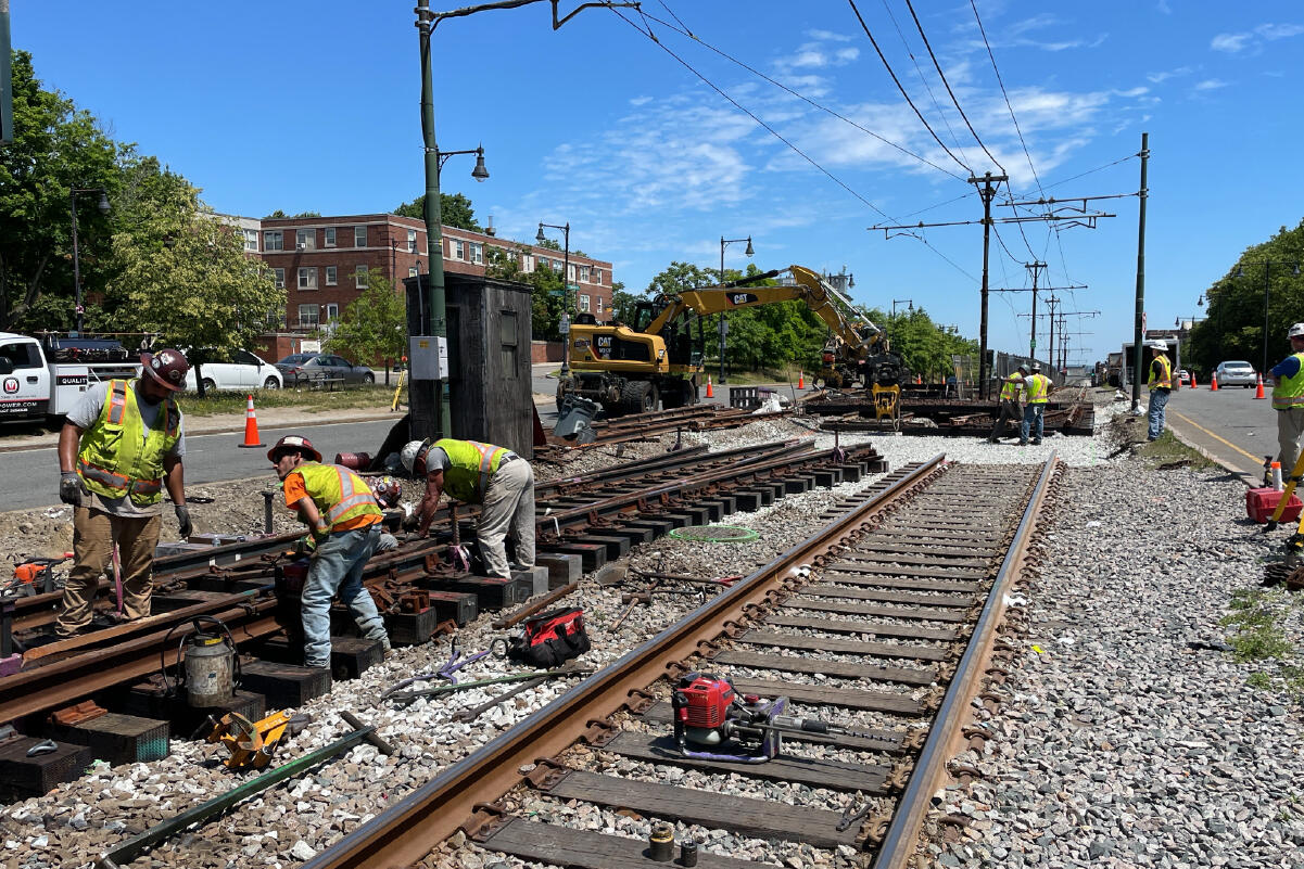 three crew members in the foreground appear to be talking with each other while they work on train tracks, with various power tools in their hands and on the tracks nearby