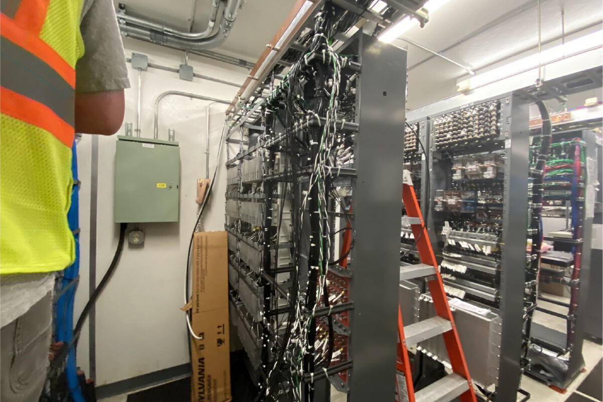 several tall racks of wires and equipment inside the signal control building, with a crew member's elbow and half of their back in the foreground