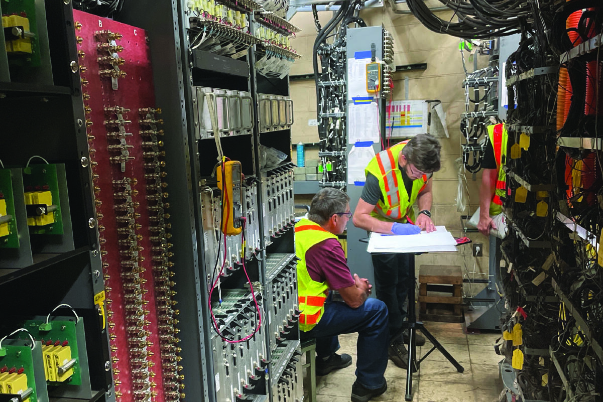 two men wearing reflective vests talking and writing on paper in a narrow room of signals and wires and switches