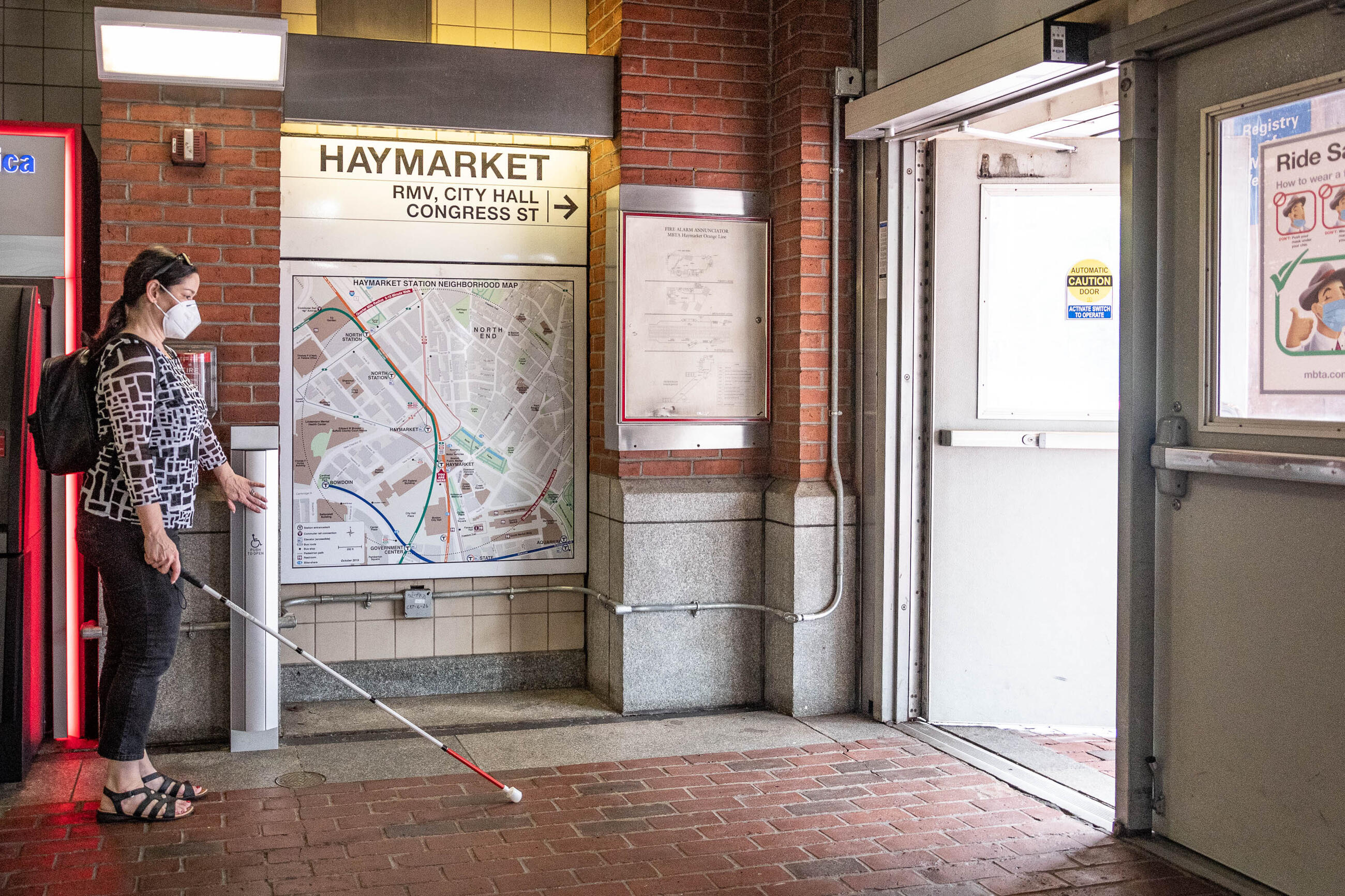 a person wearing sandals, pants, a patterned long sleeve shirt, a small backpack, a kn95 mask, and sunglasses on their head is holding a white cane and pressing the door opener button for the door in front of them. The person is inside Haymarket station at the entrance. The button is very long and can be pressed from hip height all the way down to the floor.