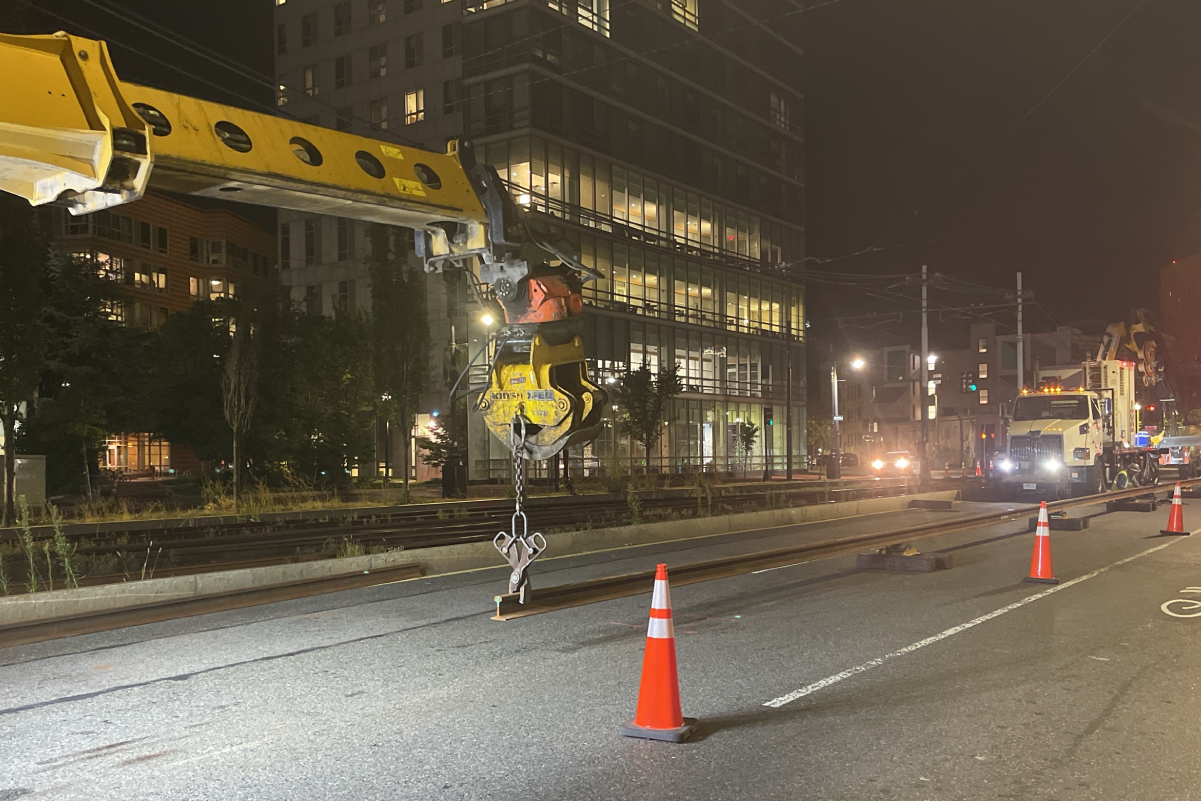 At night, a construction vehicle with a clamp holds a train track rail in place while a worker prepares the rail for installation on the Green Line E Branch.