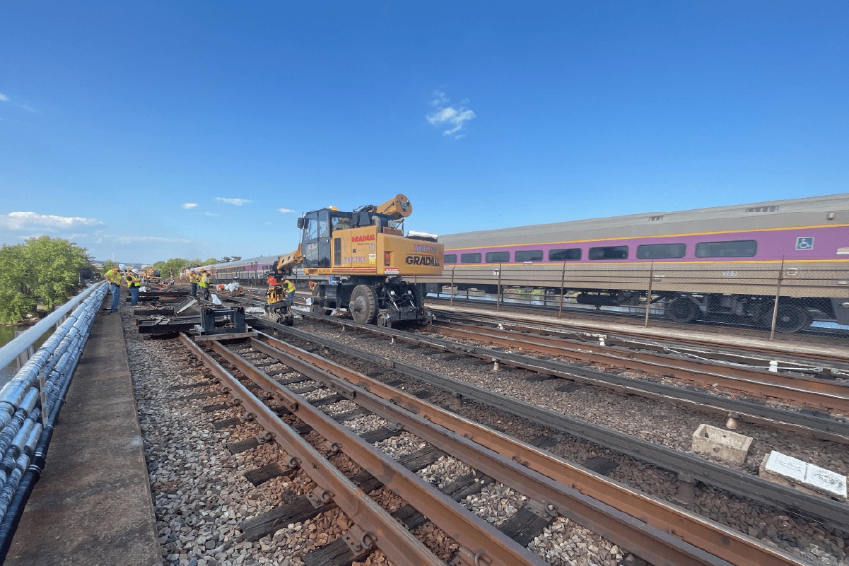 crews using big machinery and other equipment on rails on a bridge, with a commuter rail train parked on the rails behind them