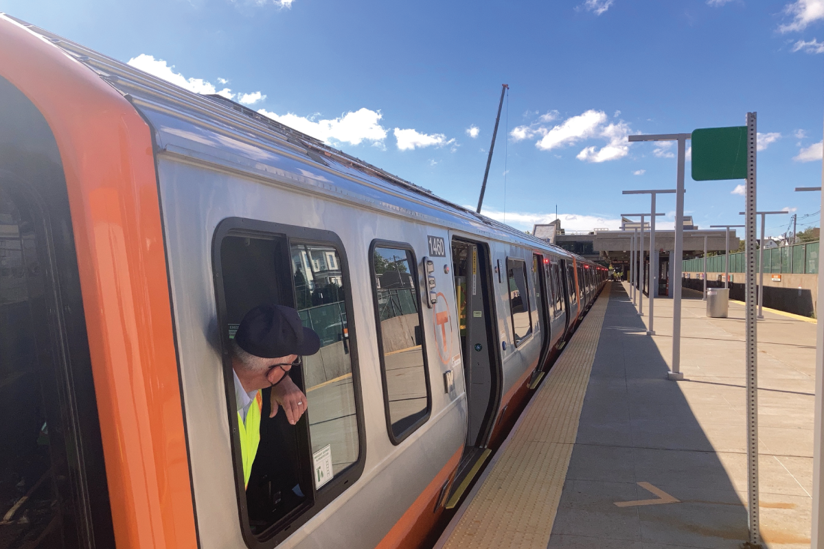 A worker looks out of the window of a new Orange Line car away from the camera and down the track outside under a blue, partly cloudy sky.