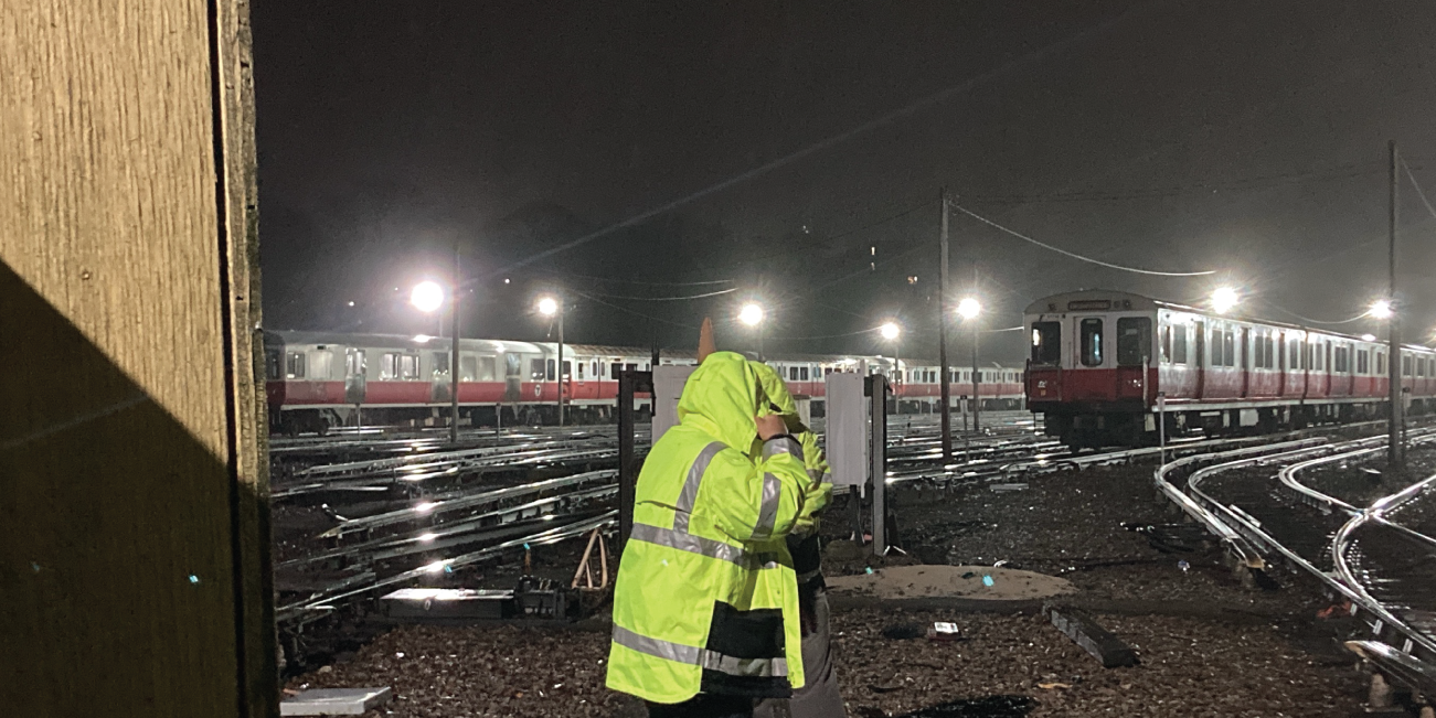 a person in a reflective rain jacket in front of many red line trains in a train yard. it's dark and rainy