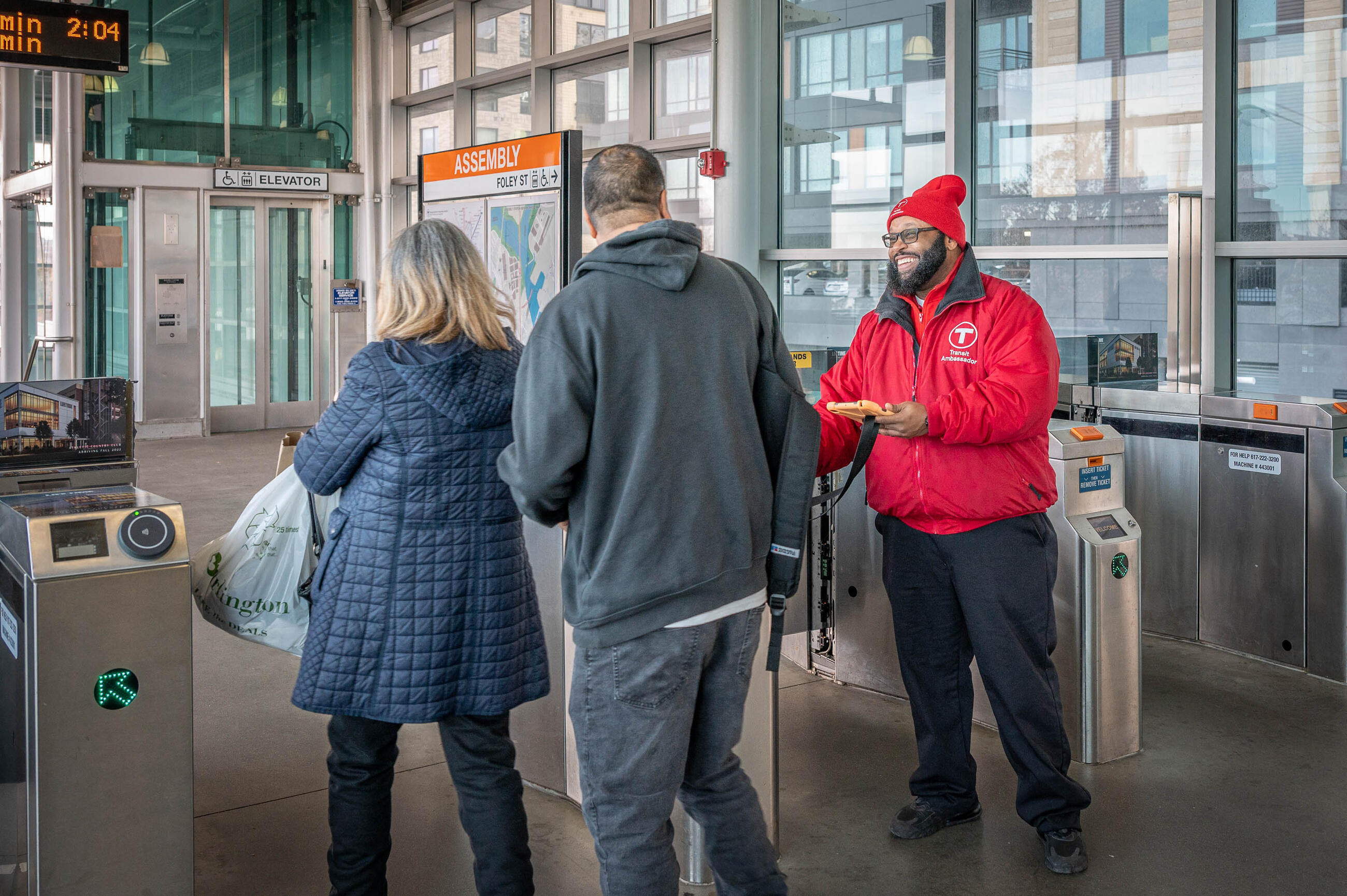 Transit ambassador in red jacket helping a pair of riders at the Assembly fare gate