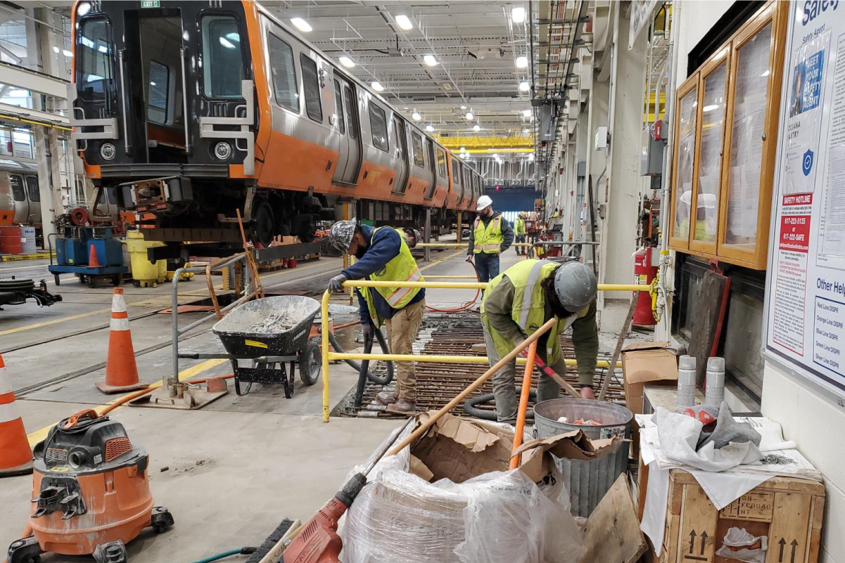 crews working on the floor inside a brightly lit facility with orange line train cars on risers behind them