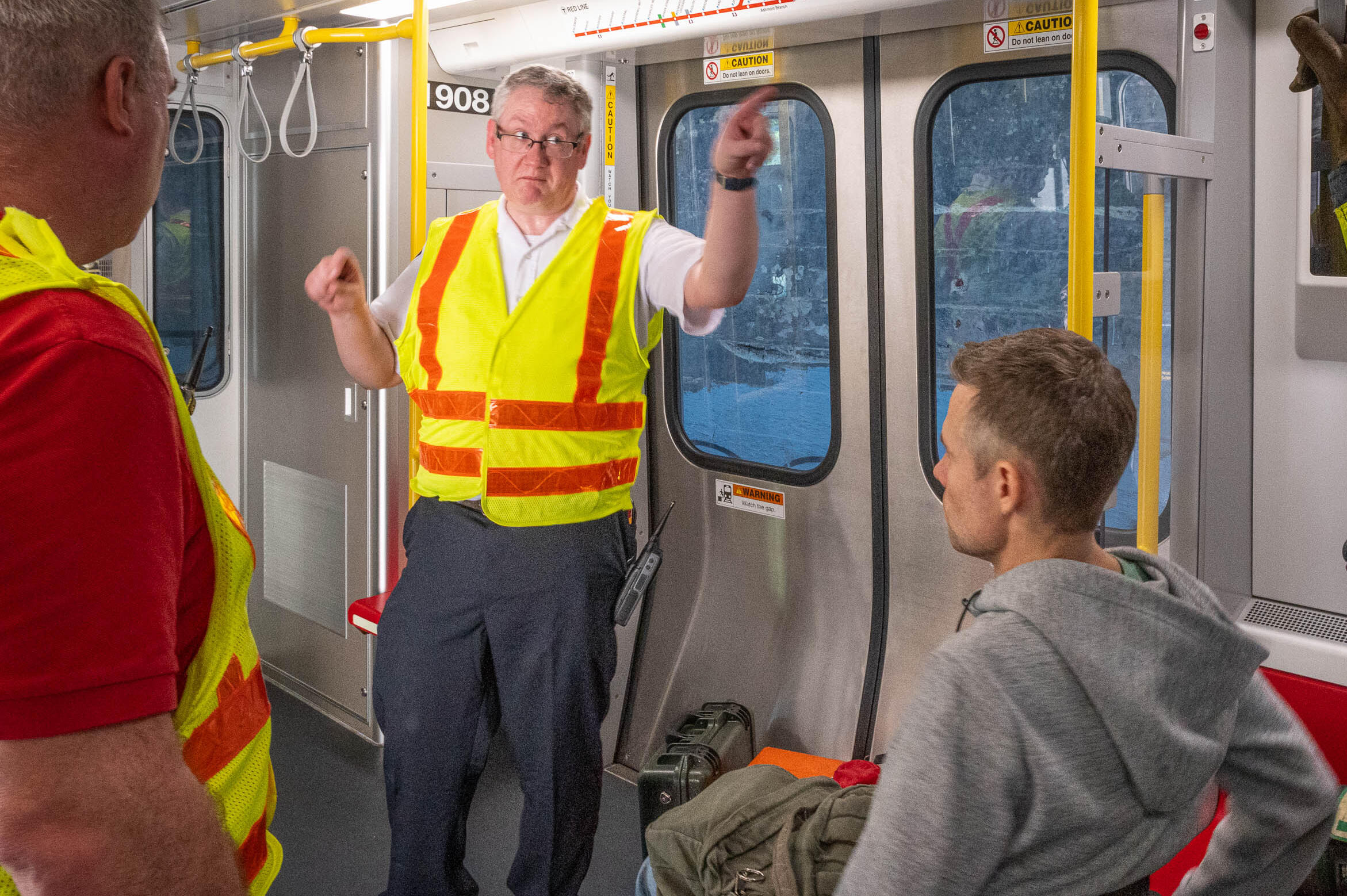 An MBTA official in a Red Line subway car is wearing a high visibility safety vest while he conducts an emergency evacuation drill. He points to the opposite end of the subway car while two trainees watch and listen to him.