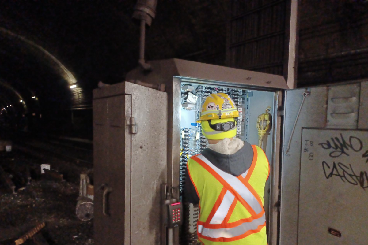 crew worker inside dark subway tunnel looking at box full of wires under a floodlight