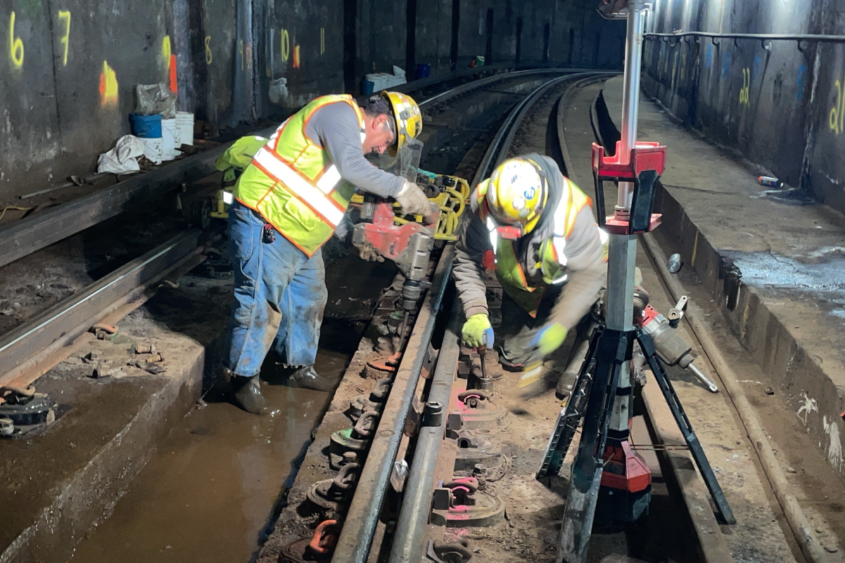 two crew members in hard hats and reflective vests using tools on train tracks under floodlights in a dark tunnel