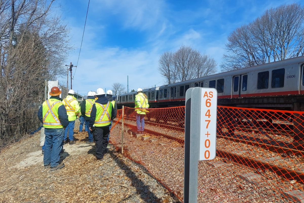 a photo from behind of seven people in coats hard hats and reflective vests standing near train tracks