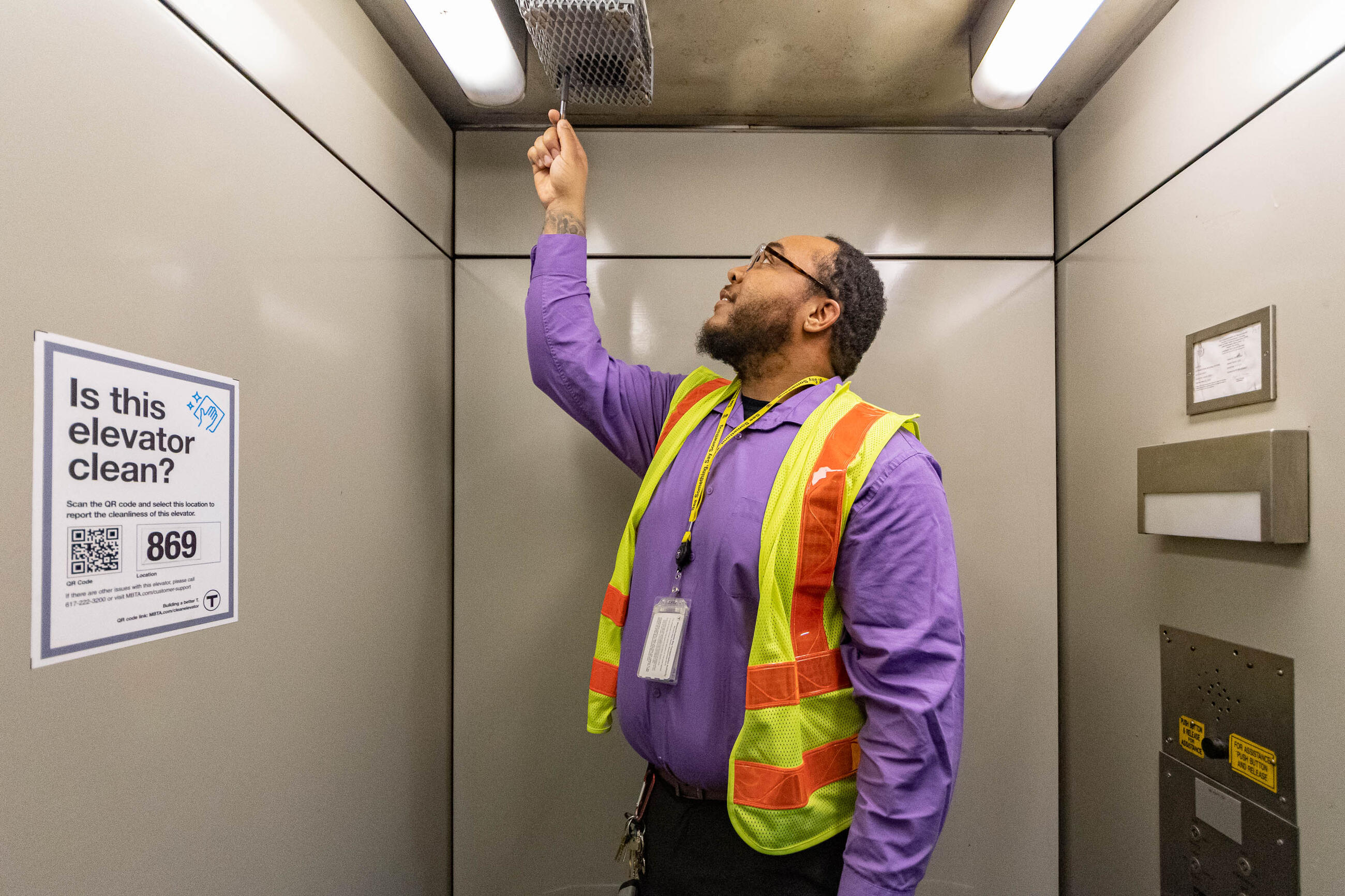 an employee in a reflective vest using a small hand tool on a urine detector sensor on the ceiling on an elevator