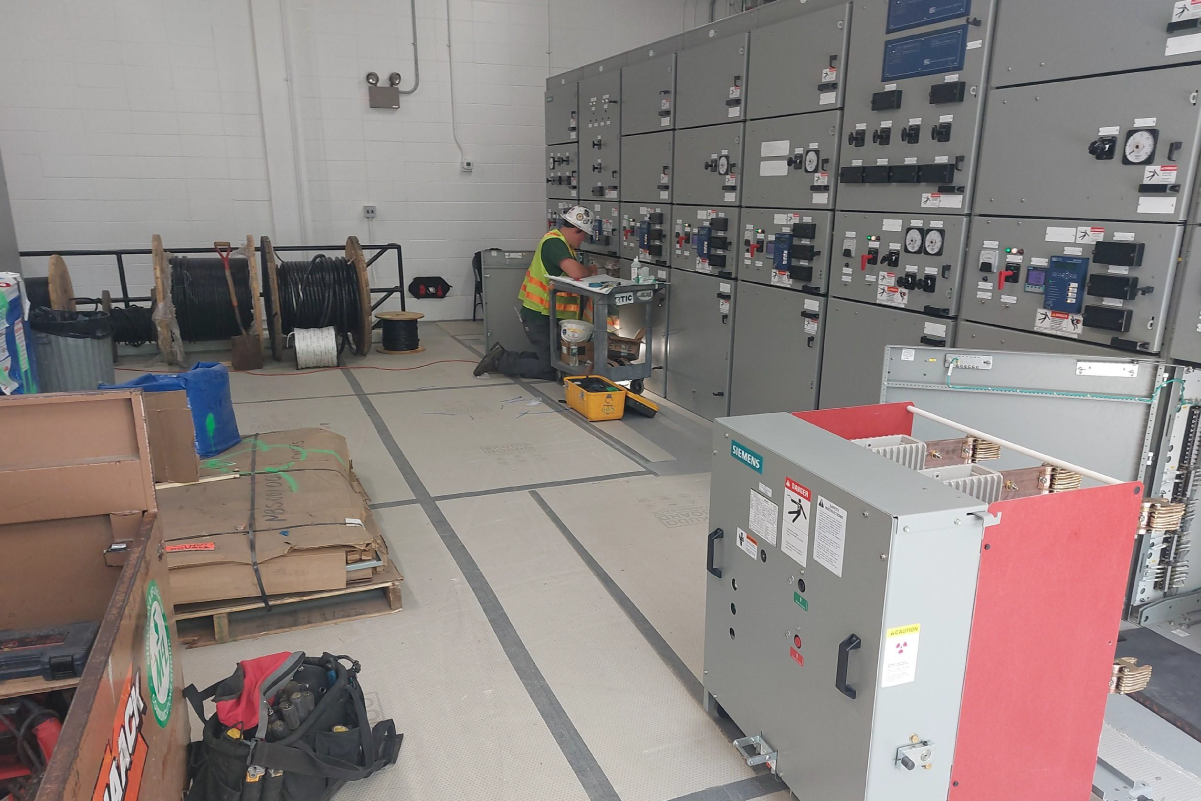 crew member kneeling on floor in front of wall of grey steel panels with various dials and switches