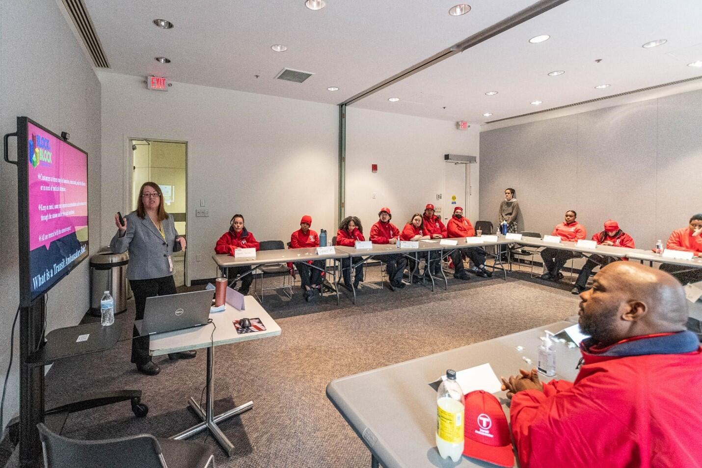 A group of people wearing red uniform shirts sit in a semi-circle facing a presentation