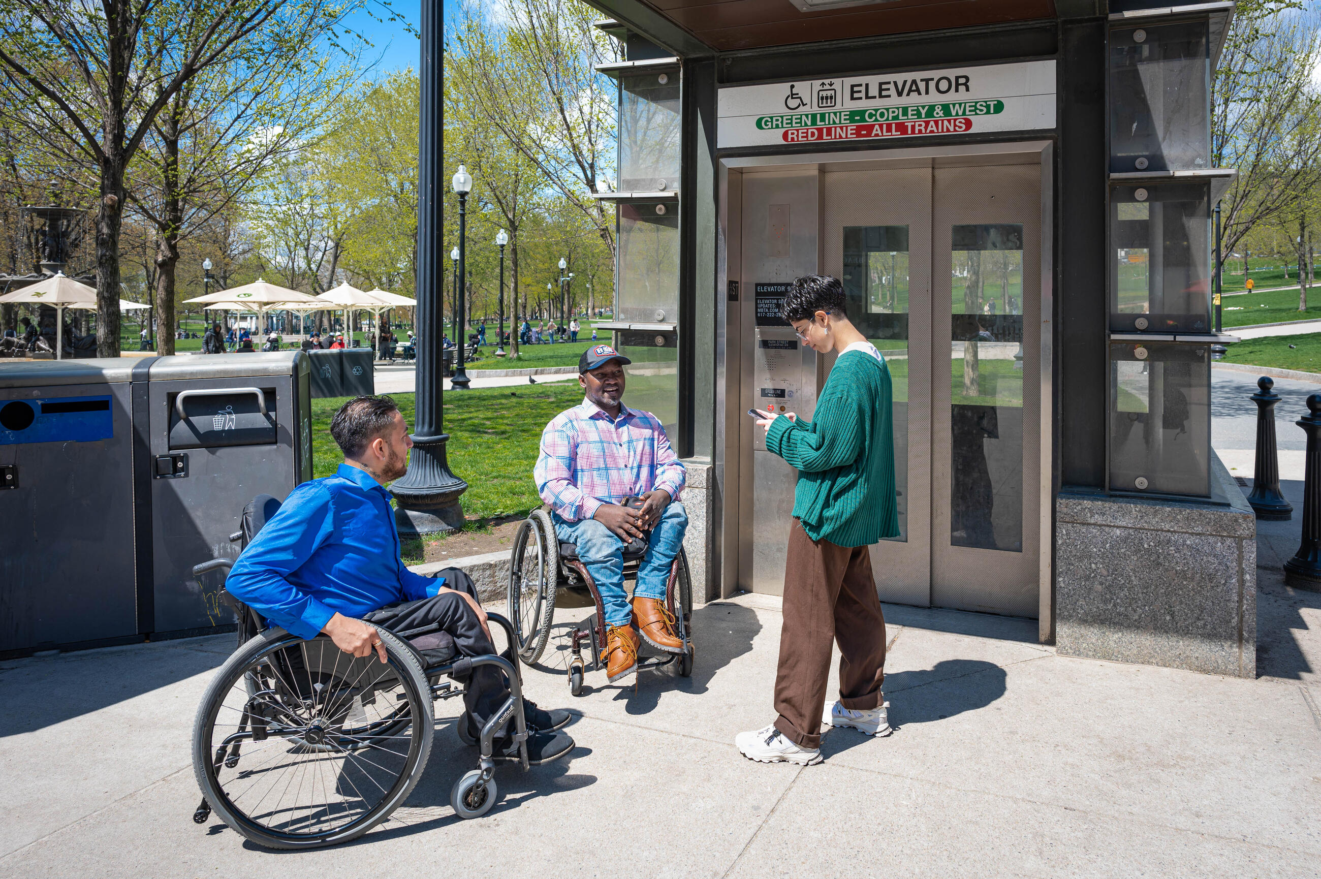 Three people are seen outside an elevator on the Boston Common. An African American man using a wheeled mobility device, a Latino man using a wheeled mobility device, and a non-binary white person using their phone to get elevator alerts. 